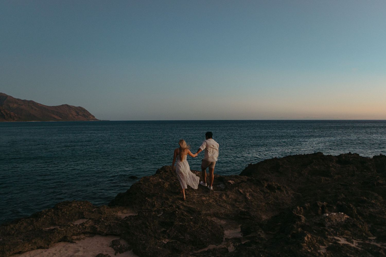 Couple standing on rocky coastal cliffs watching a serene ocean sunset with mountains in the background.
