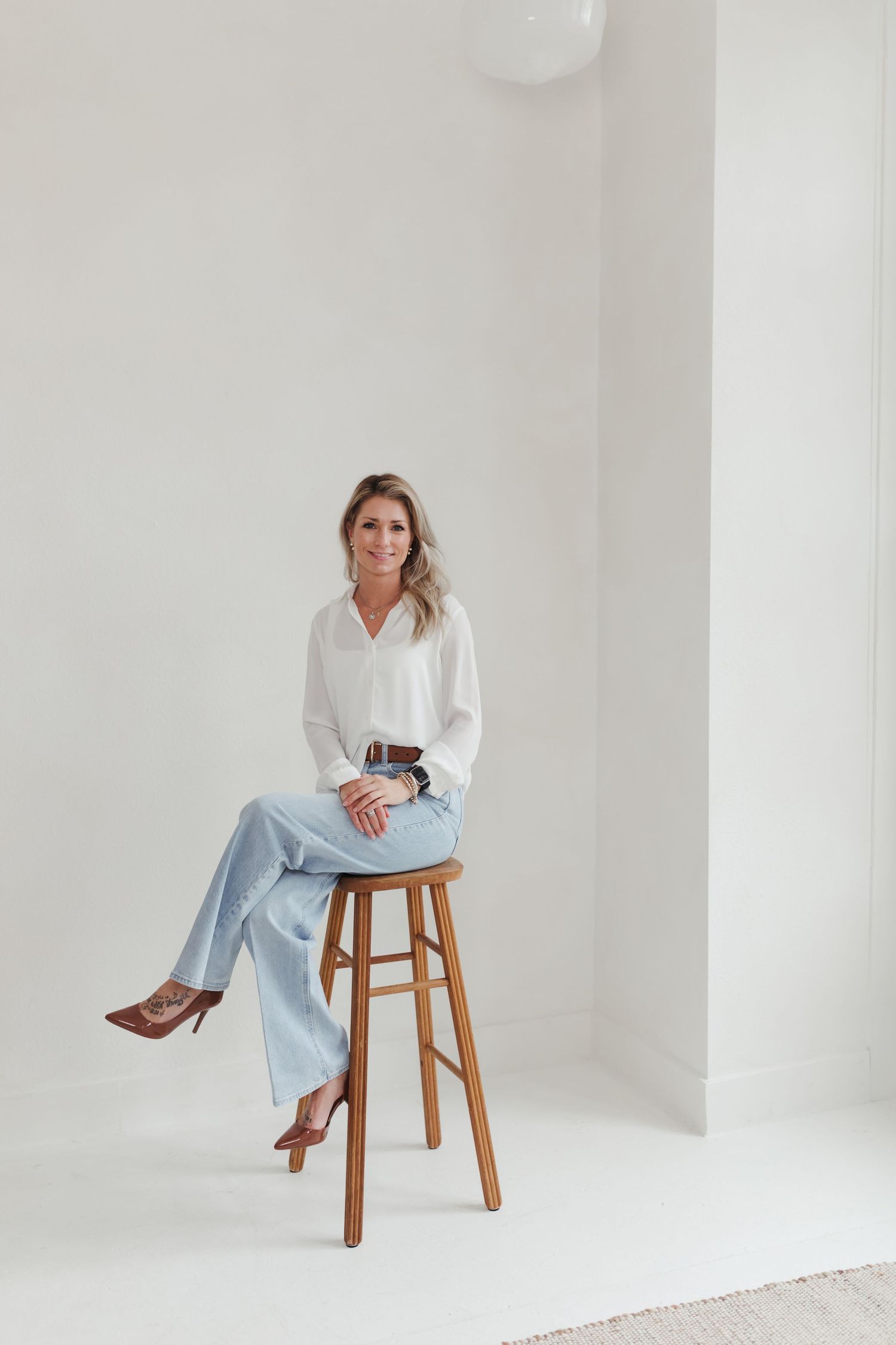 A person in a white shirt and light blue pants poses elegantly on a wooden stool against a white studio backdrop.