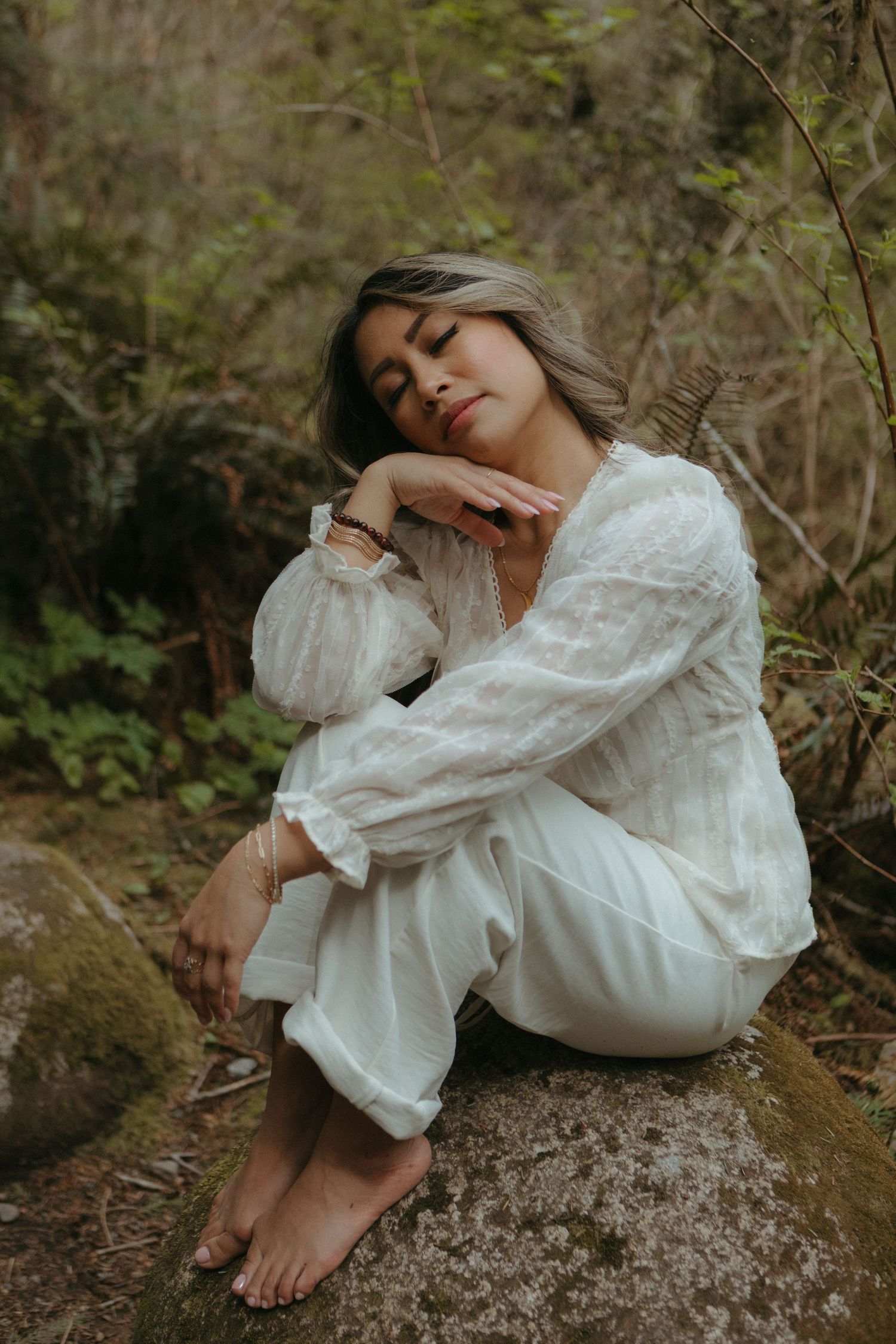 A person in a flowing white dress reclines on mossy rocks in a dreamy woodland setting.