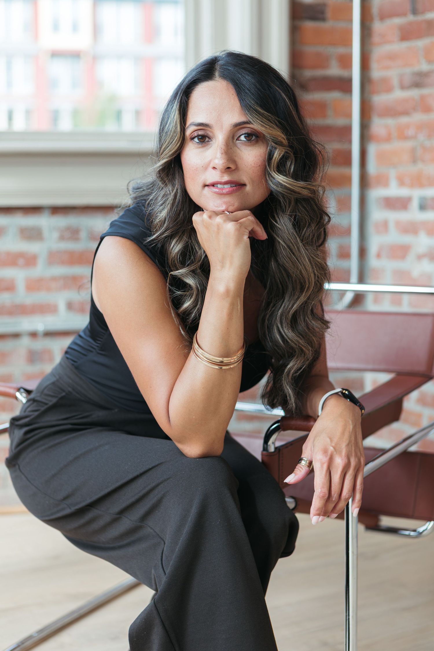 Woman with long wavy hair rests her chin on her hand while seated in a chrome chair in a bright room with exposed brick walls.