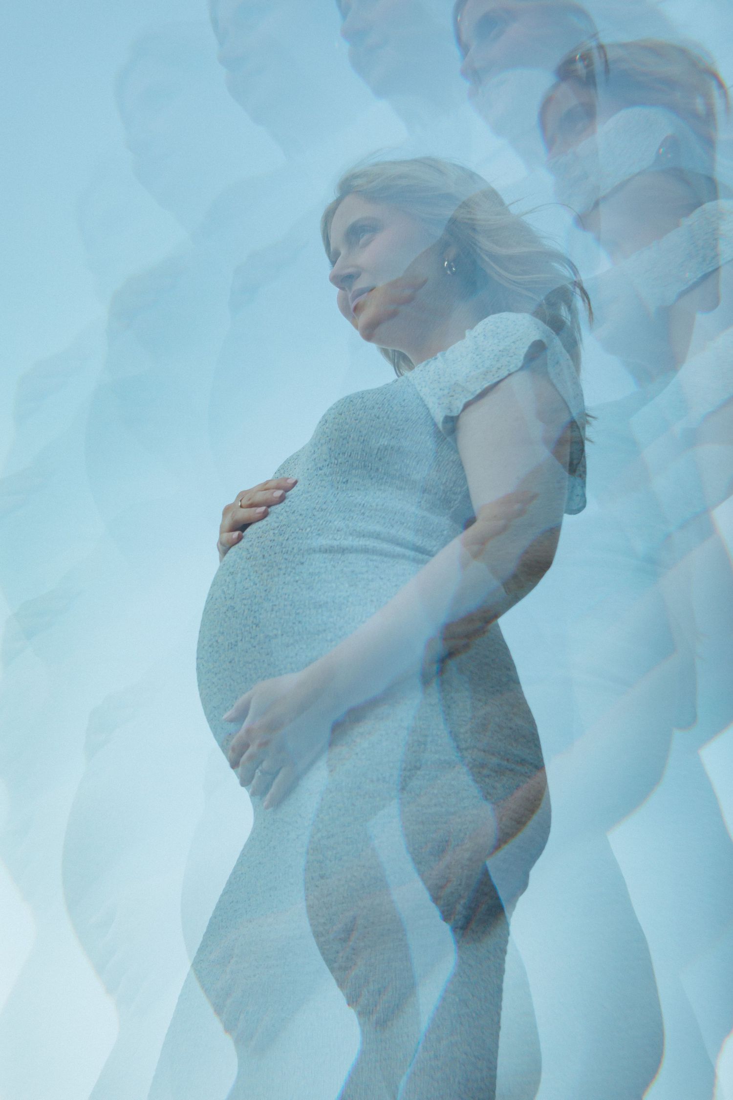 Multiple exposure photograph of a pregnant woman in a light blue dress showing her growing belly against a bright background.