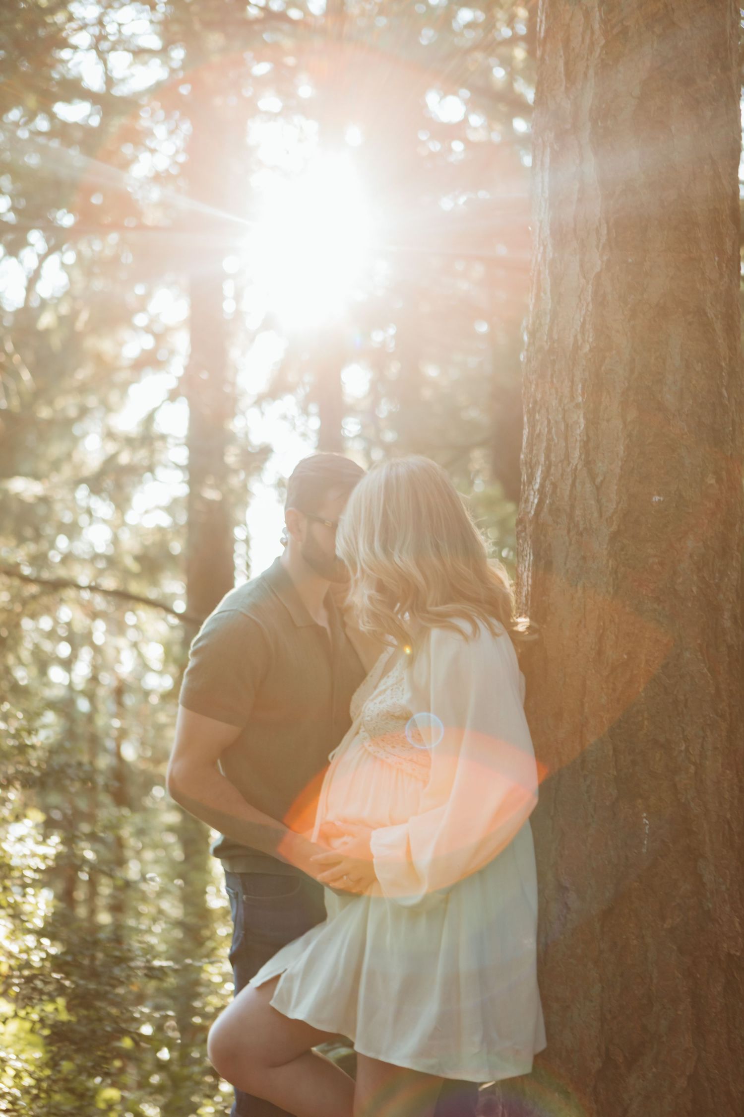 Romantic couple embraces in a sunlit forest with golden rays streaming through tall trees creating a dreamy atmosphere.