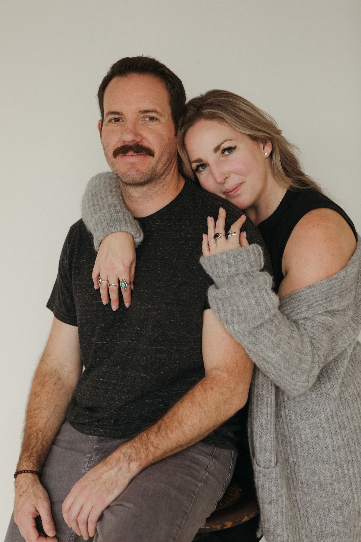 A smiling couple posing together, woman in gray cardigan leaning on man in dark t-shirt, both wearing rings.