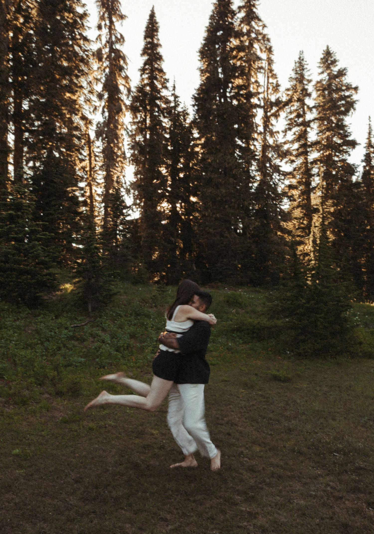 A couple embraces and spins together in a romantic dance against a backdrop of tall evergreen trees at sunset.