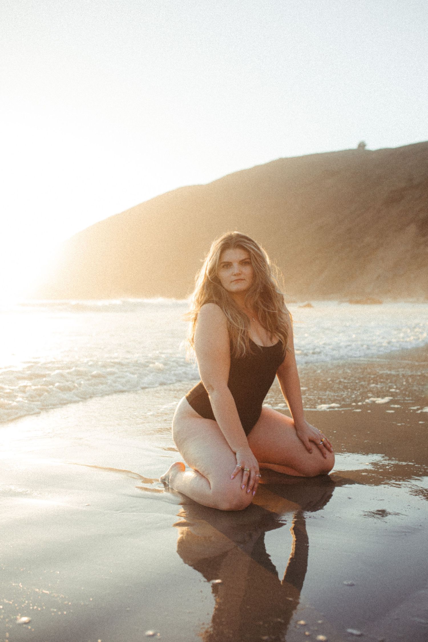 A person in a black swimsuit poses on a reflective beach at sunset with golden light and coastal cliffs in background.