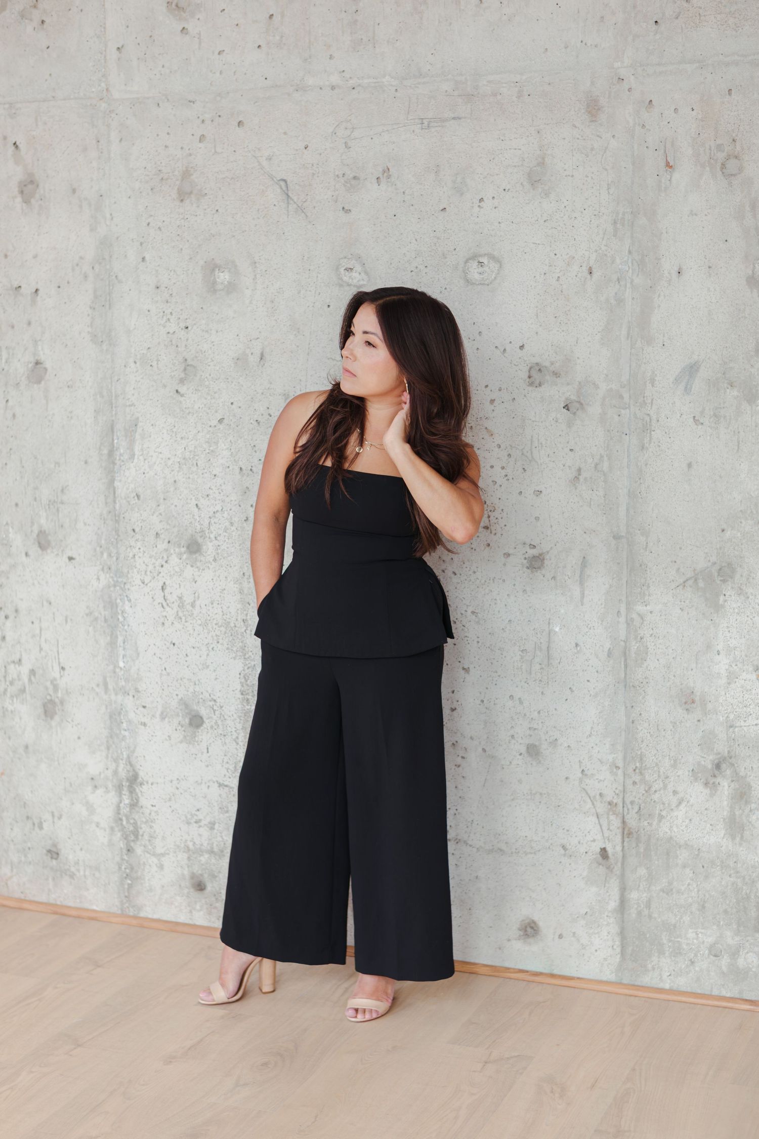 Woman with long dark hair poses against a concrete wall in a black strapless peplum top, wide-leg pants and nude block-heel sandals.