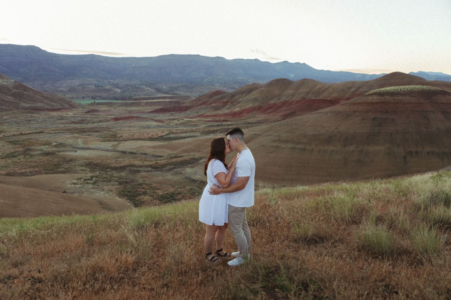 Couple embracing on a scenic overlook of the Painted Hills in Oregon at sunset, wearing white outfits.