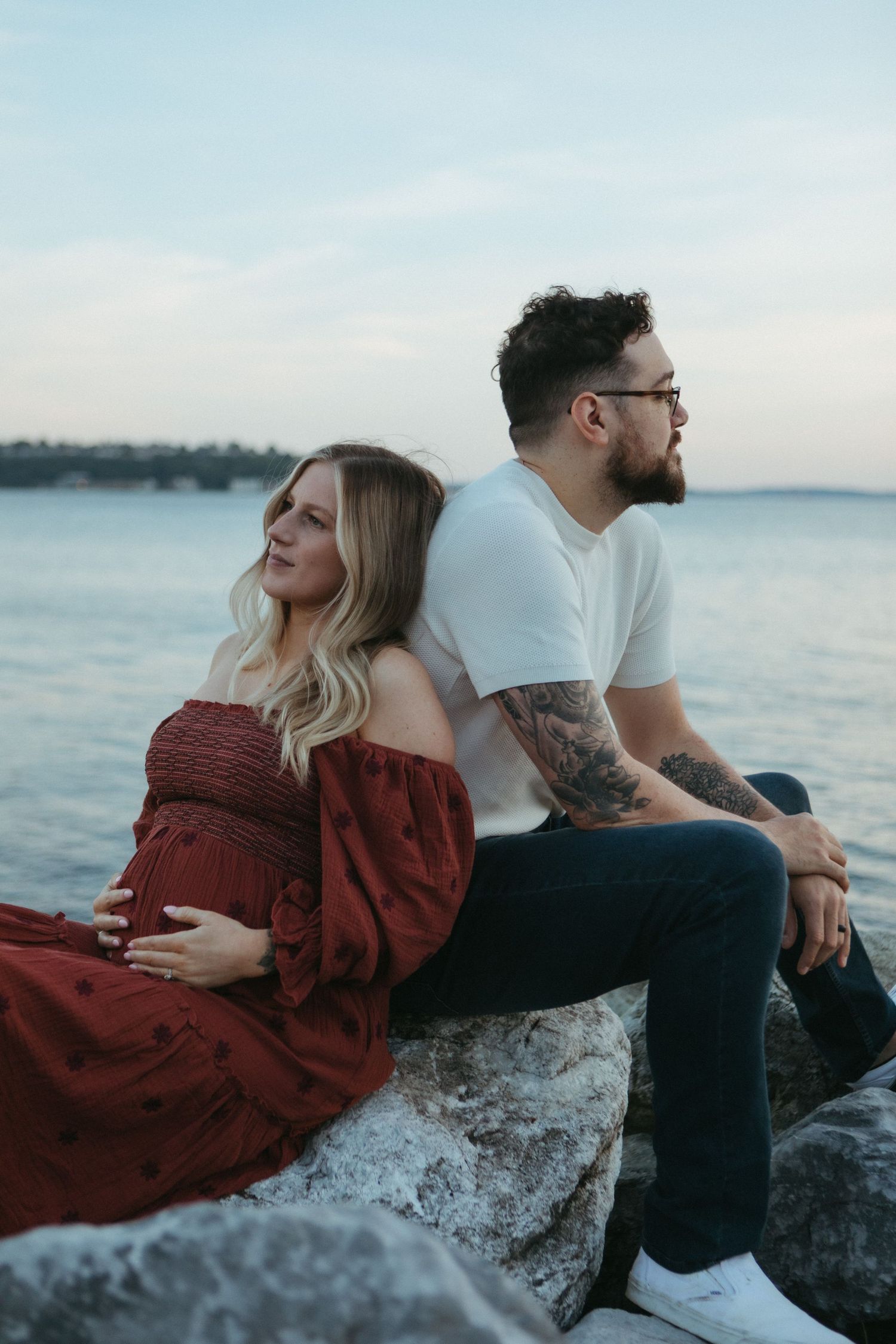 Couple sits back-to-back on coastal rocks at sunset, with her in red dress and him in white t-shirt with tattoos.