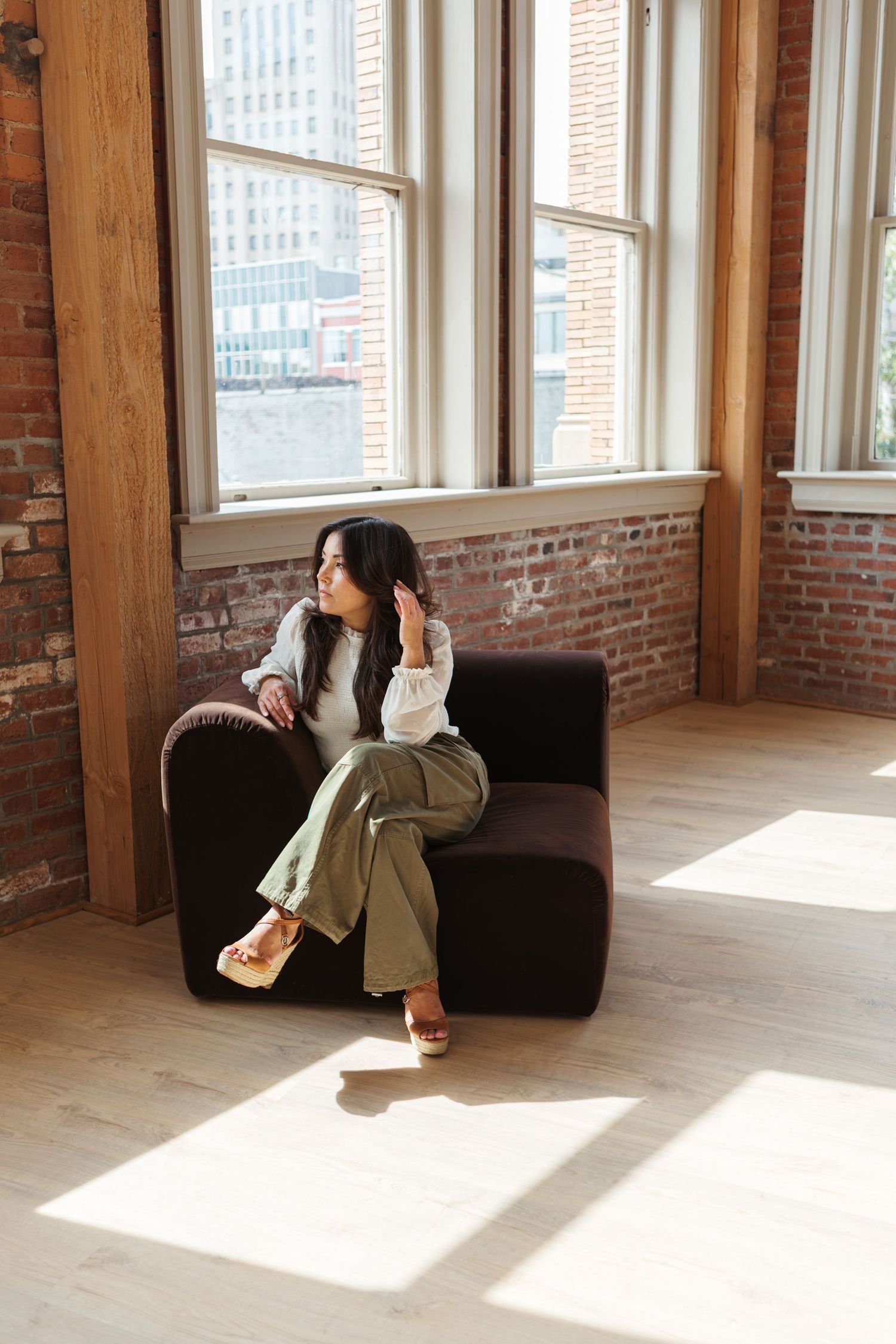 Woman with long dark hair sits on a brown velvet chair in a sunlit loft with exposed brick walls, tall windows and hardwood floors.