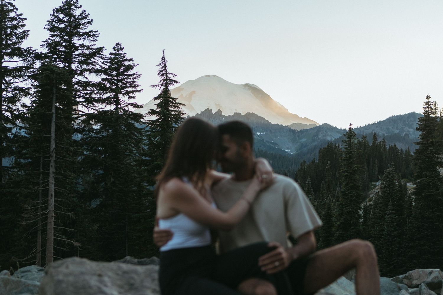 Couples embrace in a romantic mountain setting with evergreen trees and golden sunlight.
