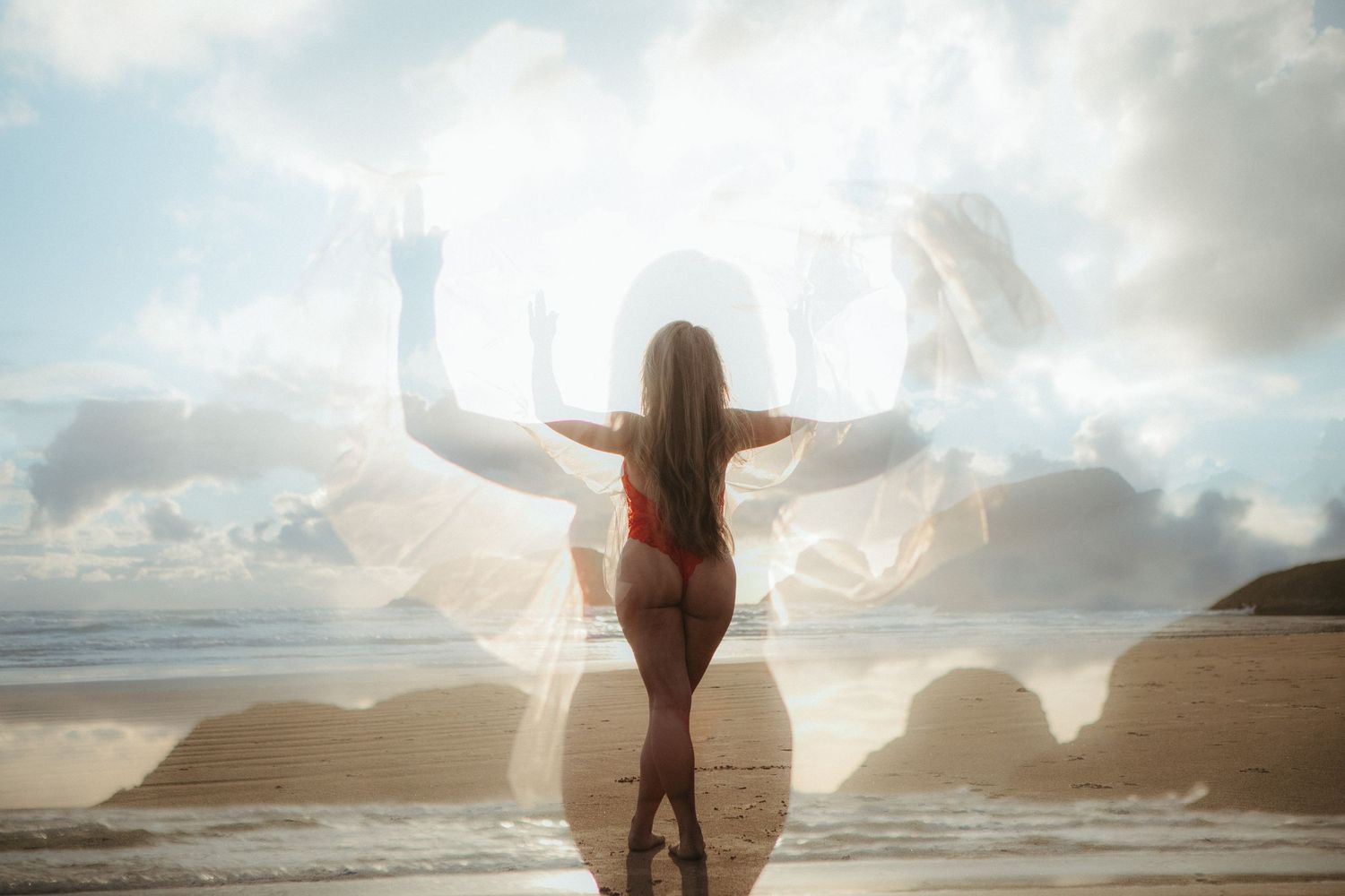 A silhouetted figure stands with outstretched arms against a dramatic cloudy sky at sunset on a beach.