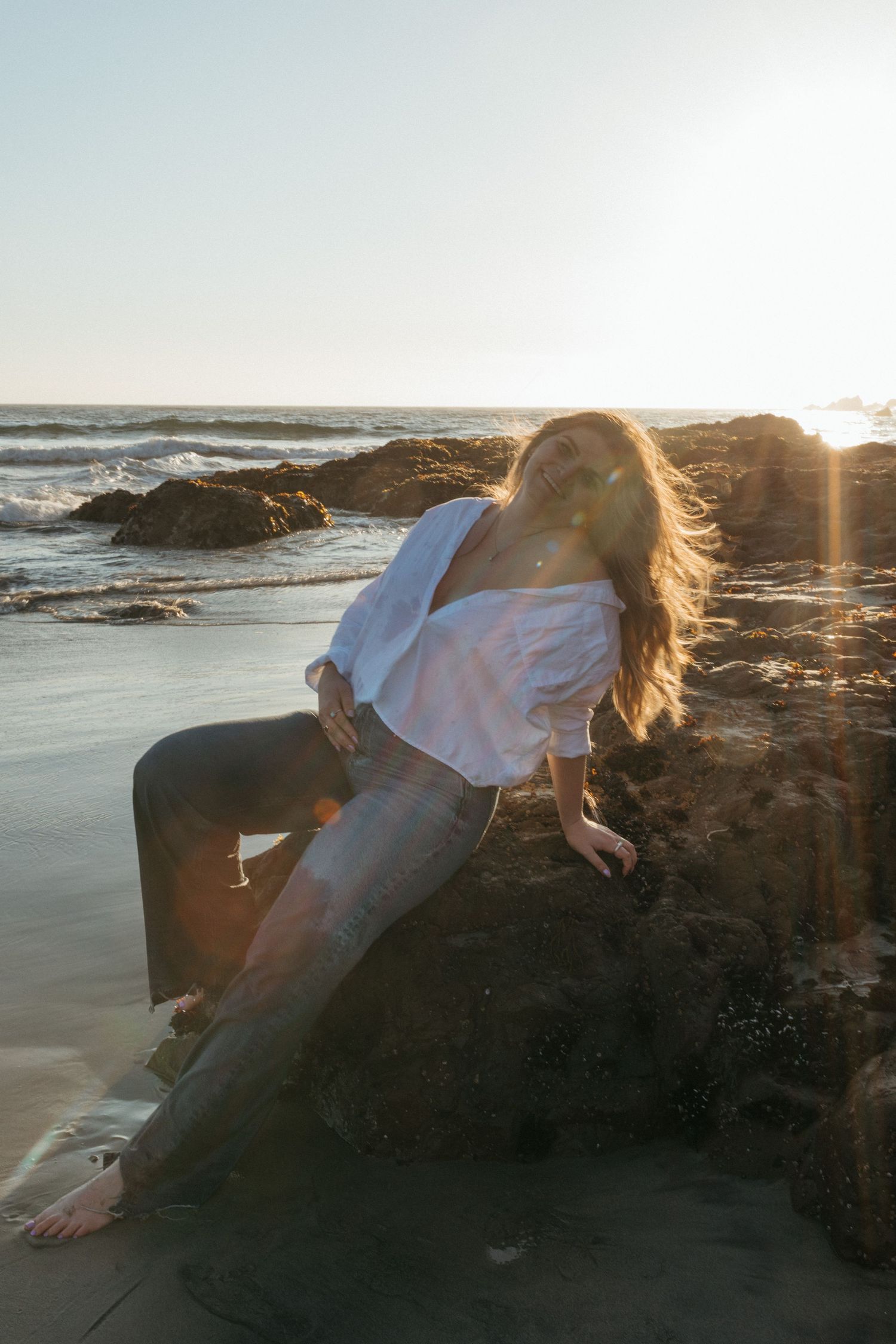 A dreamy sunset photo of someone reclining on coastal rocks as waves crash behind them.