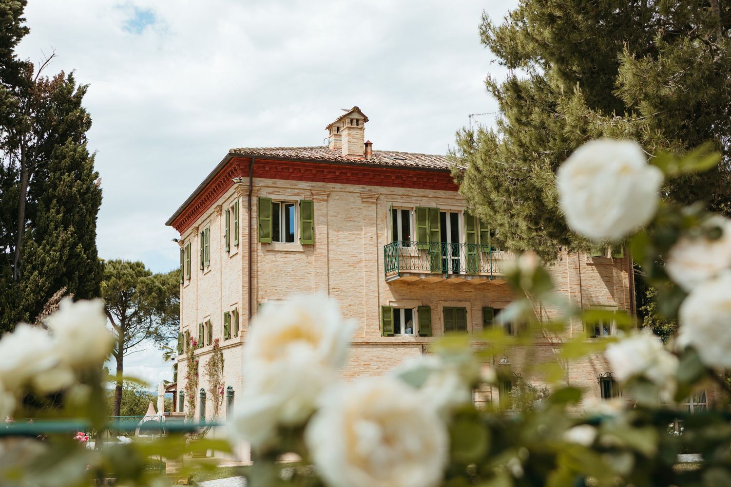 Elegant Italian villa with green shutters surrounded by white roses and cypress trees under a cloudy sky.