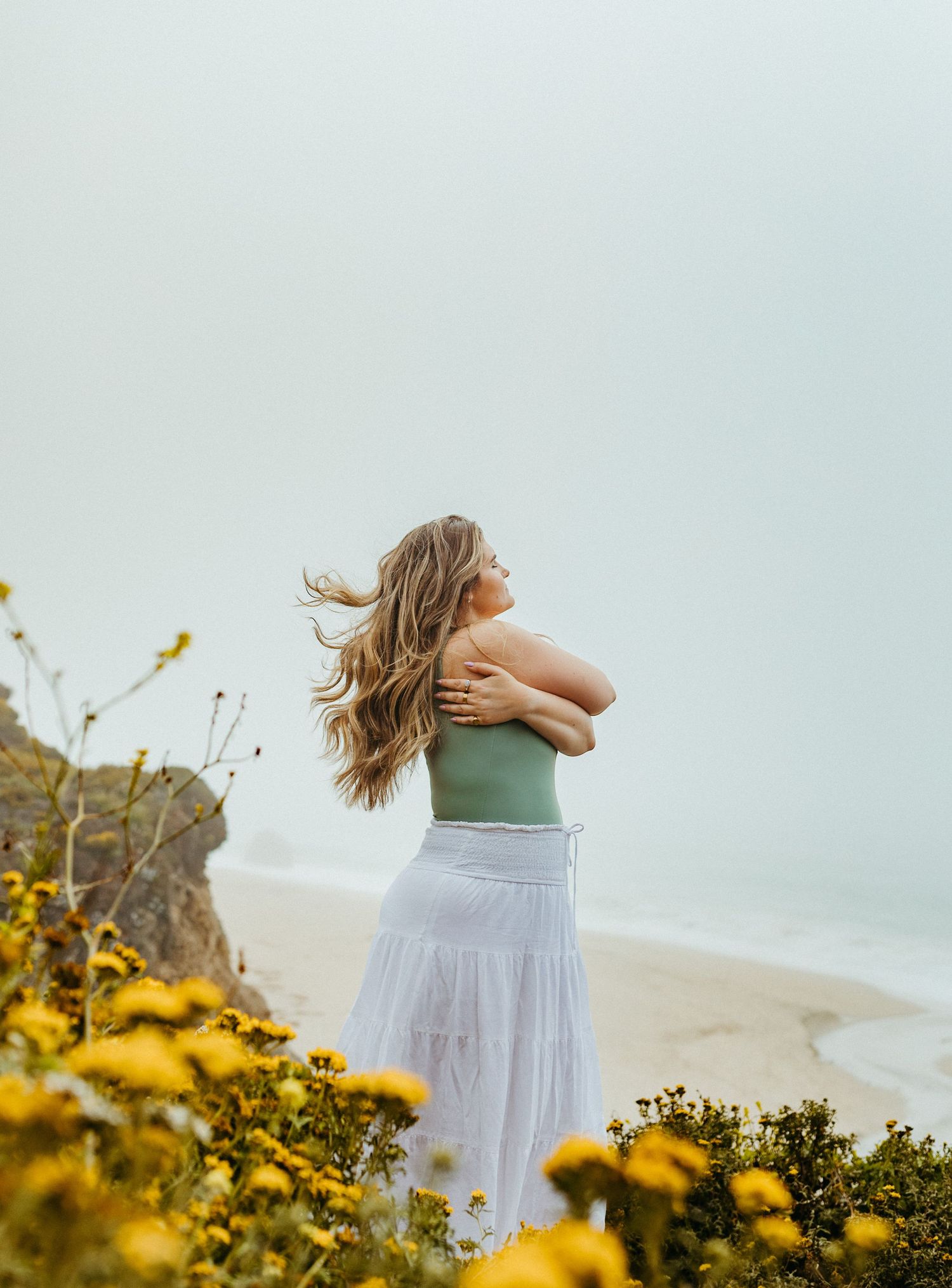 Person in white dress walking through yellow wildflowers along a coastal cliff with ocean views.