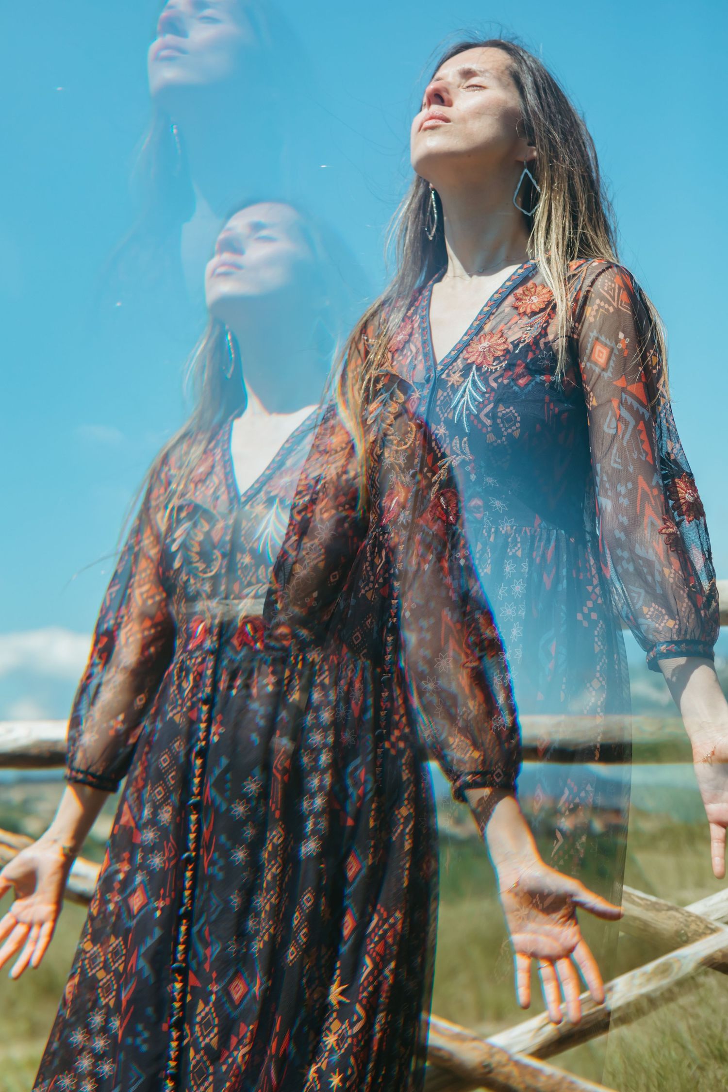 A woman in a bohemian floral dress stands outdoors with a dreamy double exposure reflection against a blue sky.