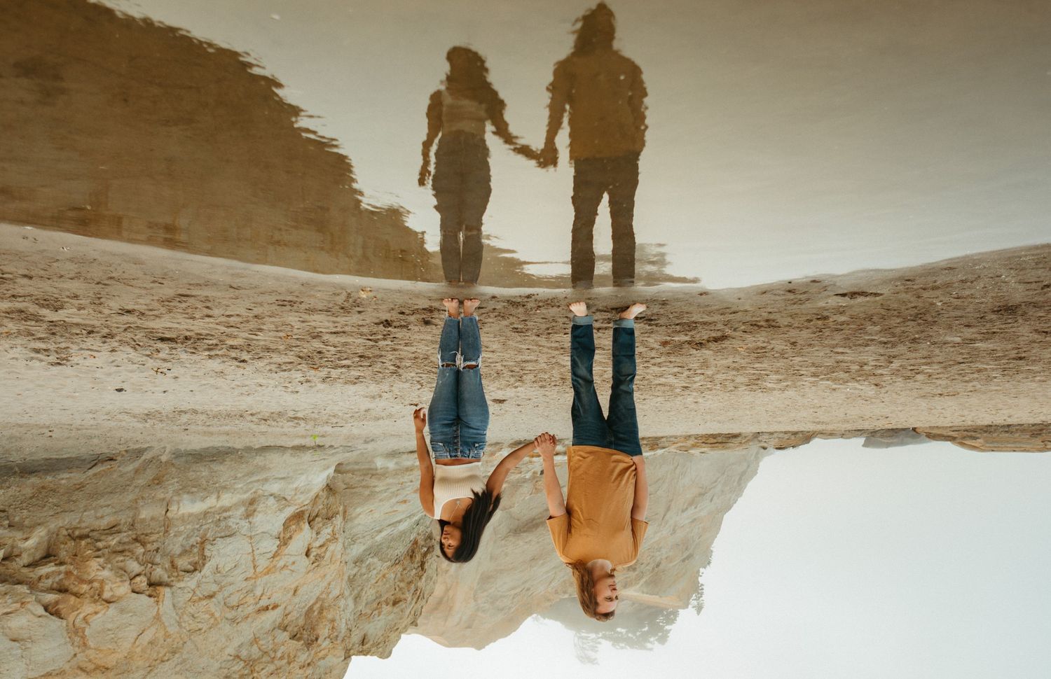 Couple holding hands reflected in a puddle on a sandy beach, creating a stunning upside-down mirror image.