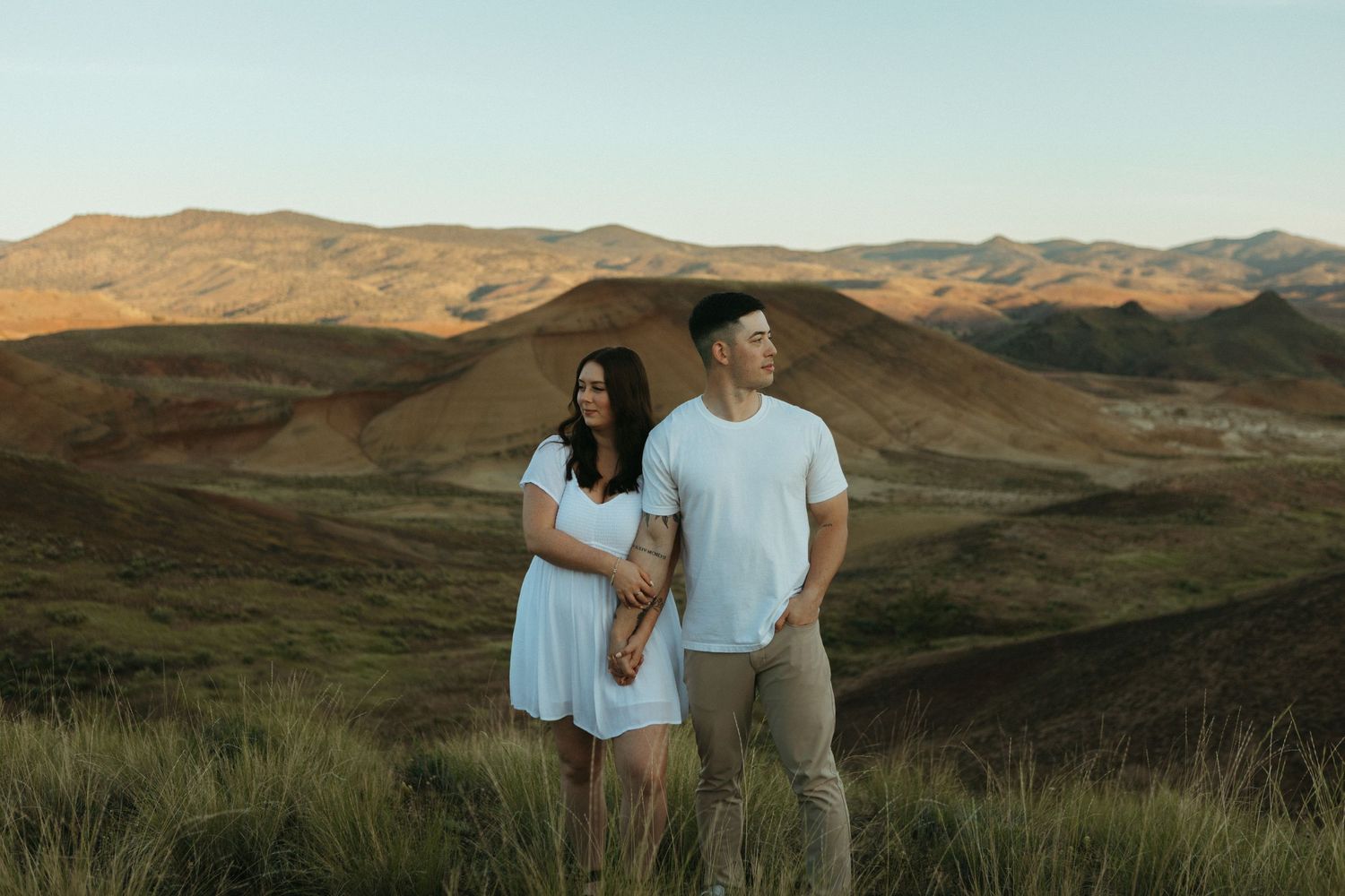 Couple standing together in white and tan outfits with scenic desert mountains at golden hour.