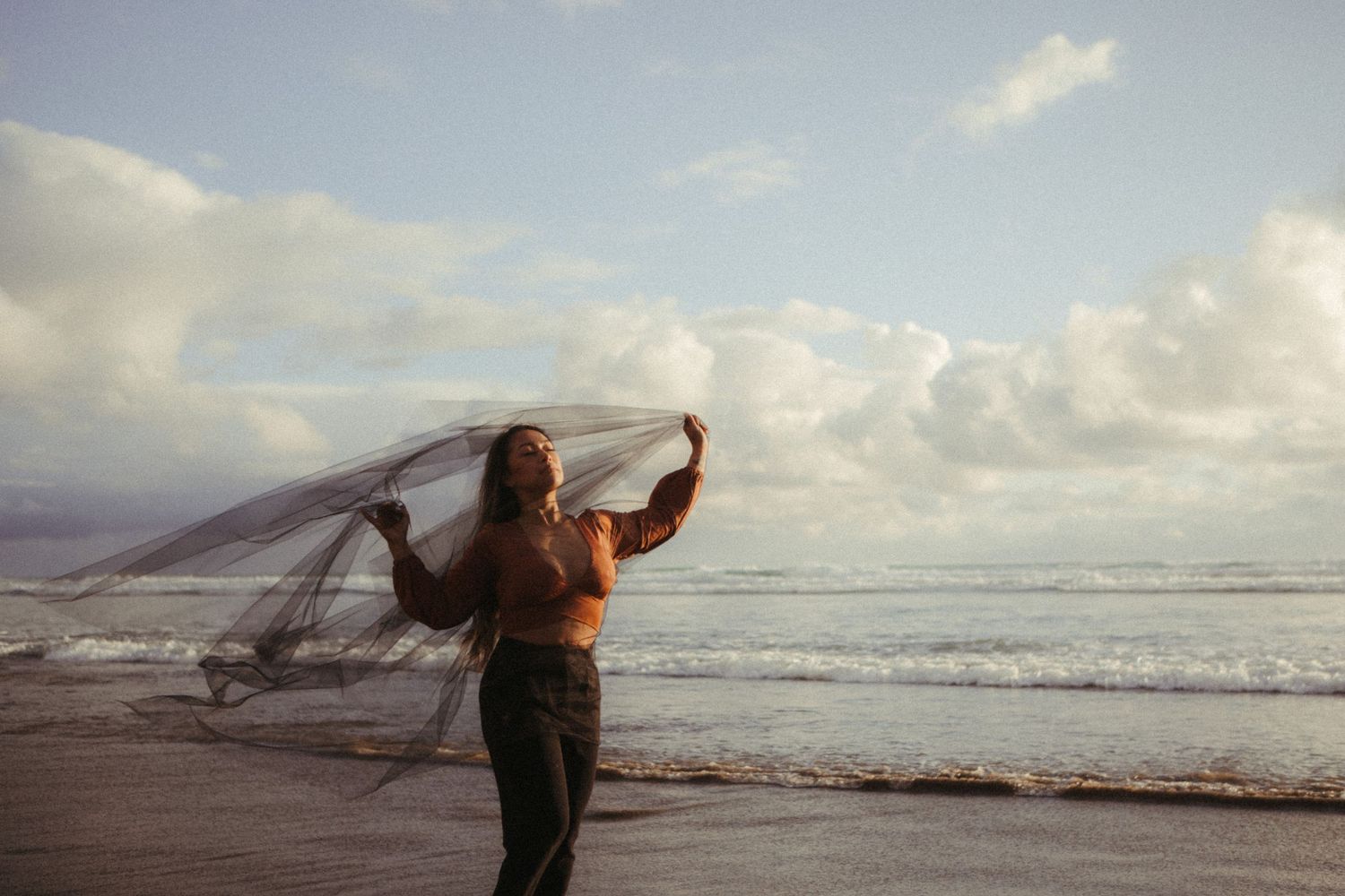 A flowing veil billows in the ocean breeze as waves crash on a scenic beach at sunset.
