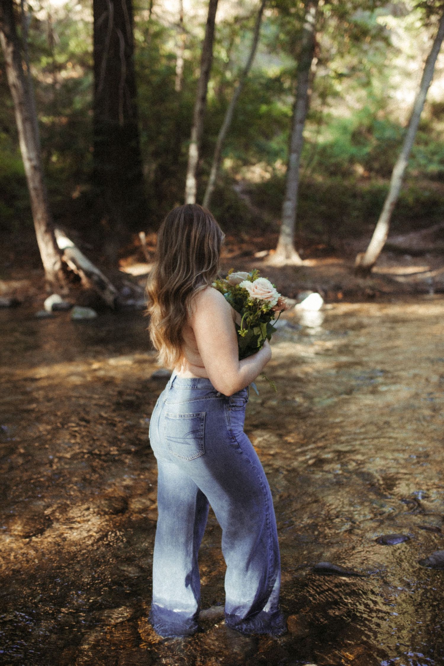 Someone in denim overalls crouches in a sunlit forest clearing surrounded by trees.