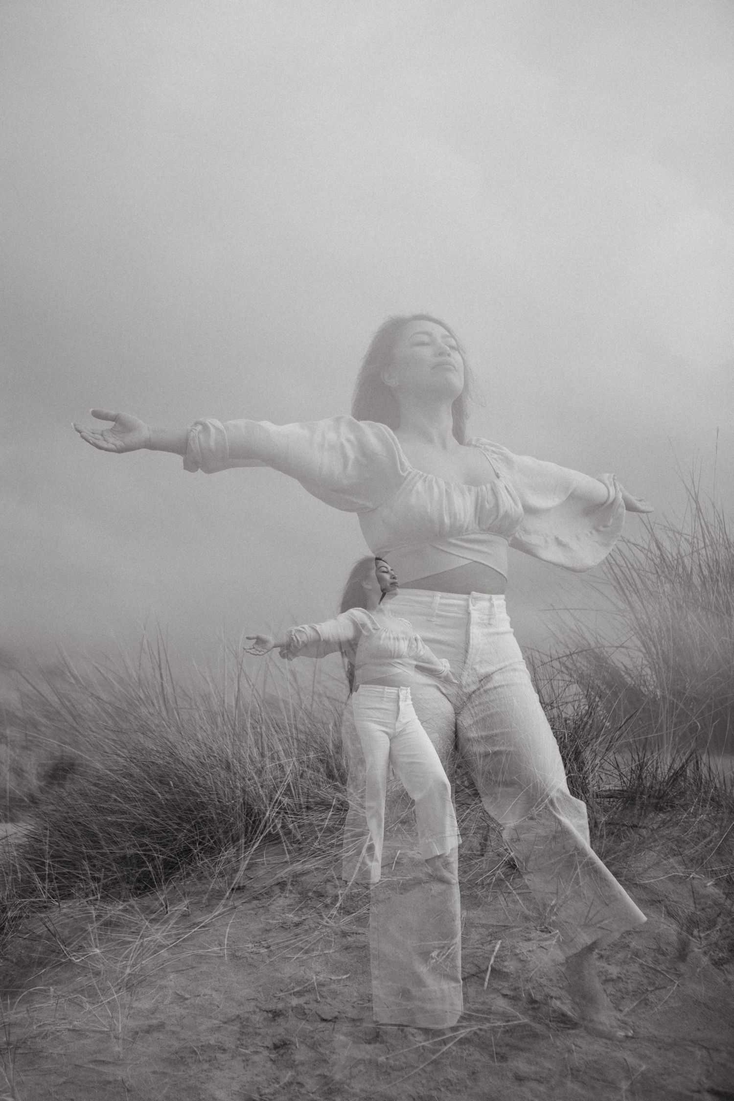 A person in white clothing stands with arms outstretched on a misty hillside in a dreamy black and white photograph.