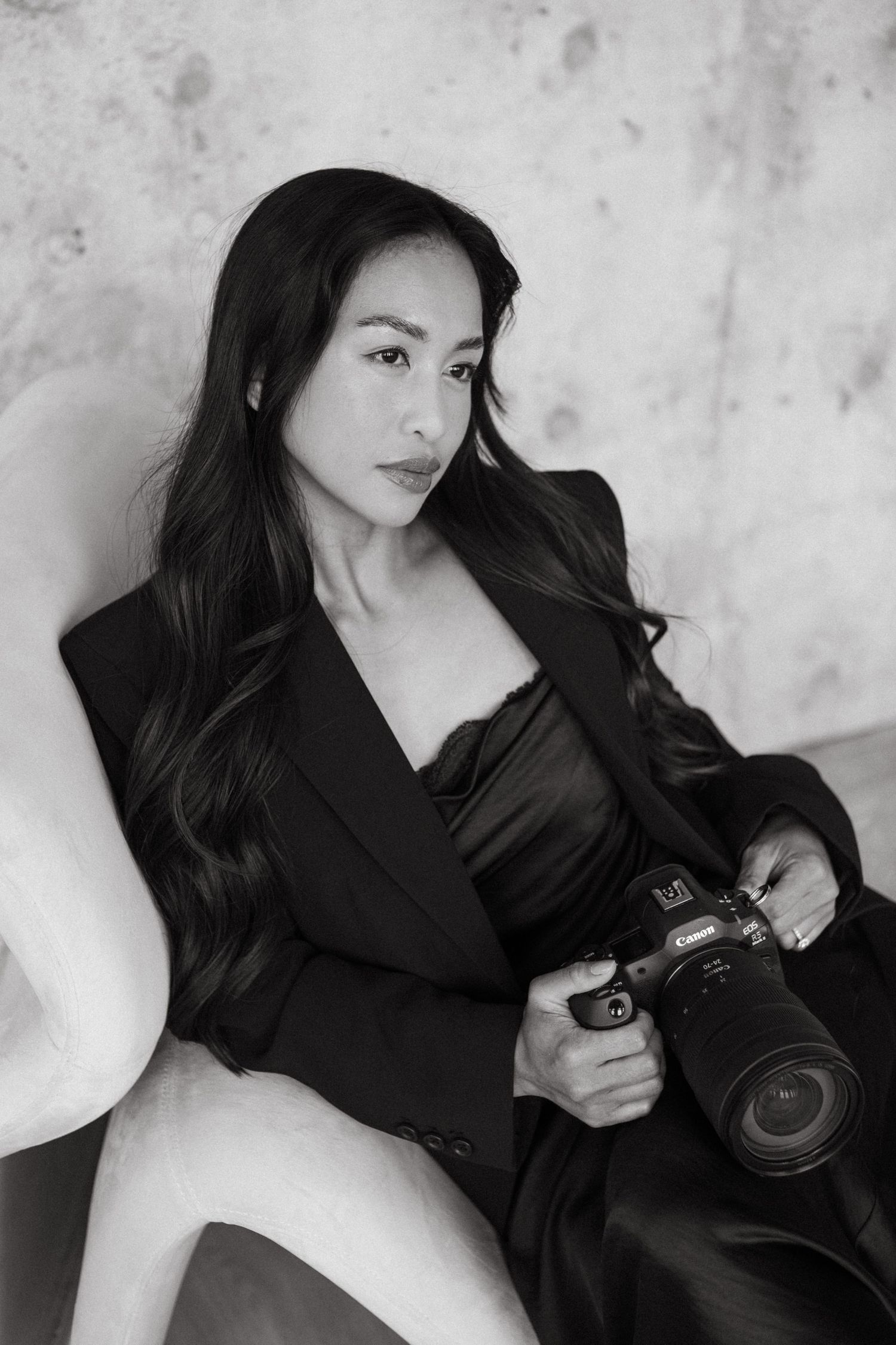 Black and white portrait of a woman with long wavy hair in a dark blazer, seated on a curved chair and holding a Canon mirrorless camera.