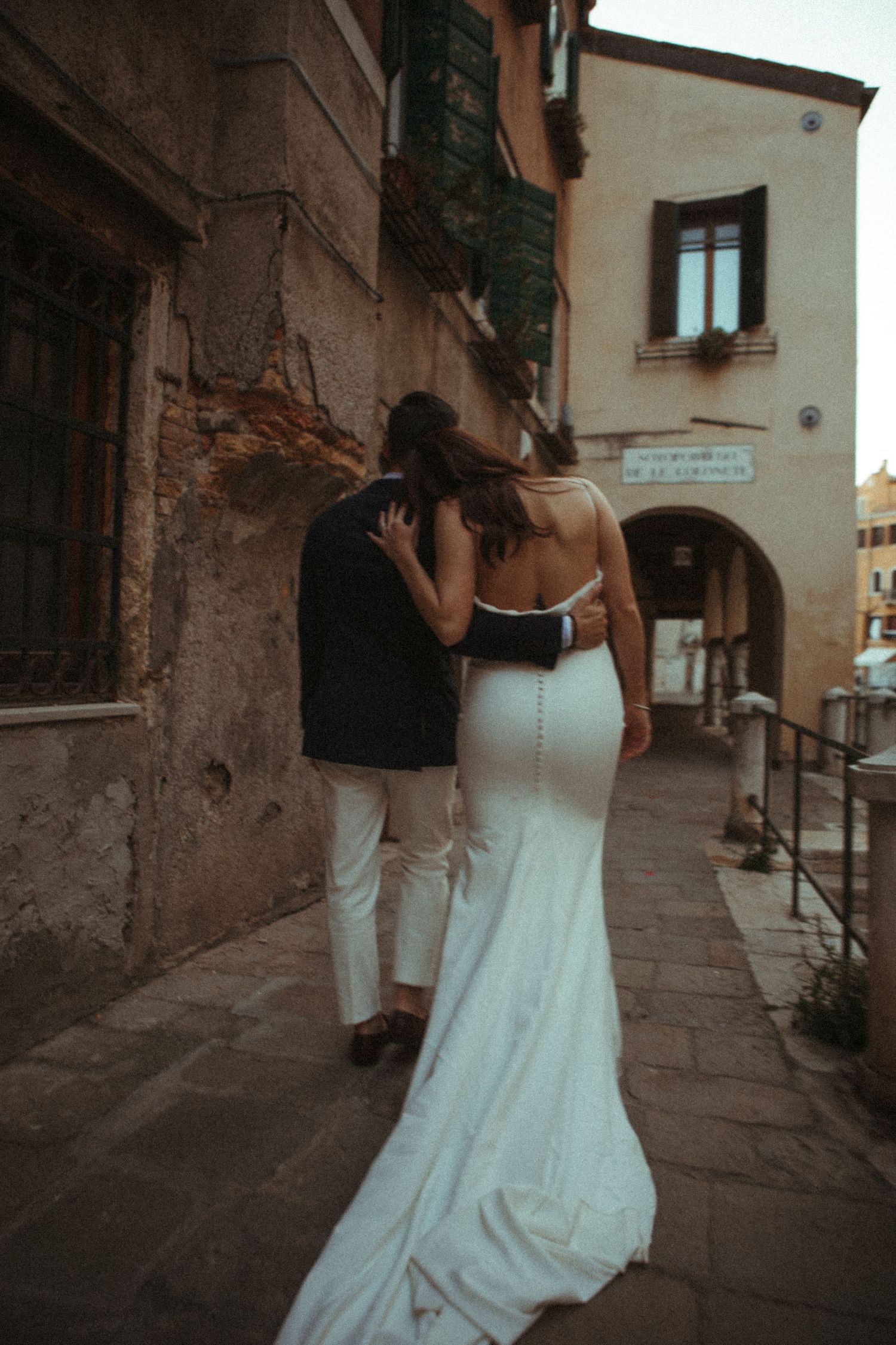 A bride and groom walk hand in hand through a charming European cobblestone street, her backless white gown trailing behind.