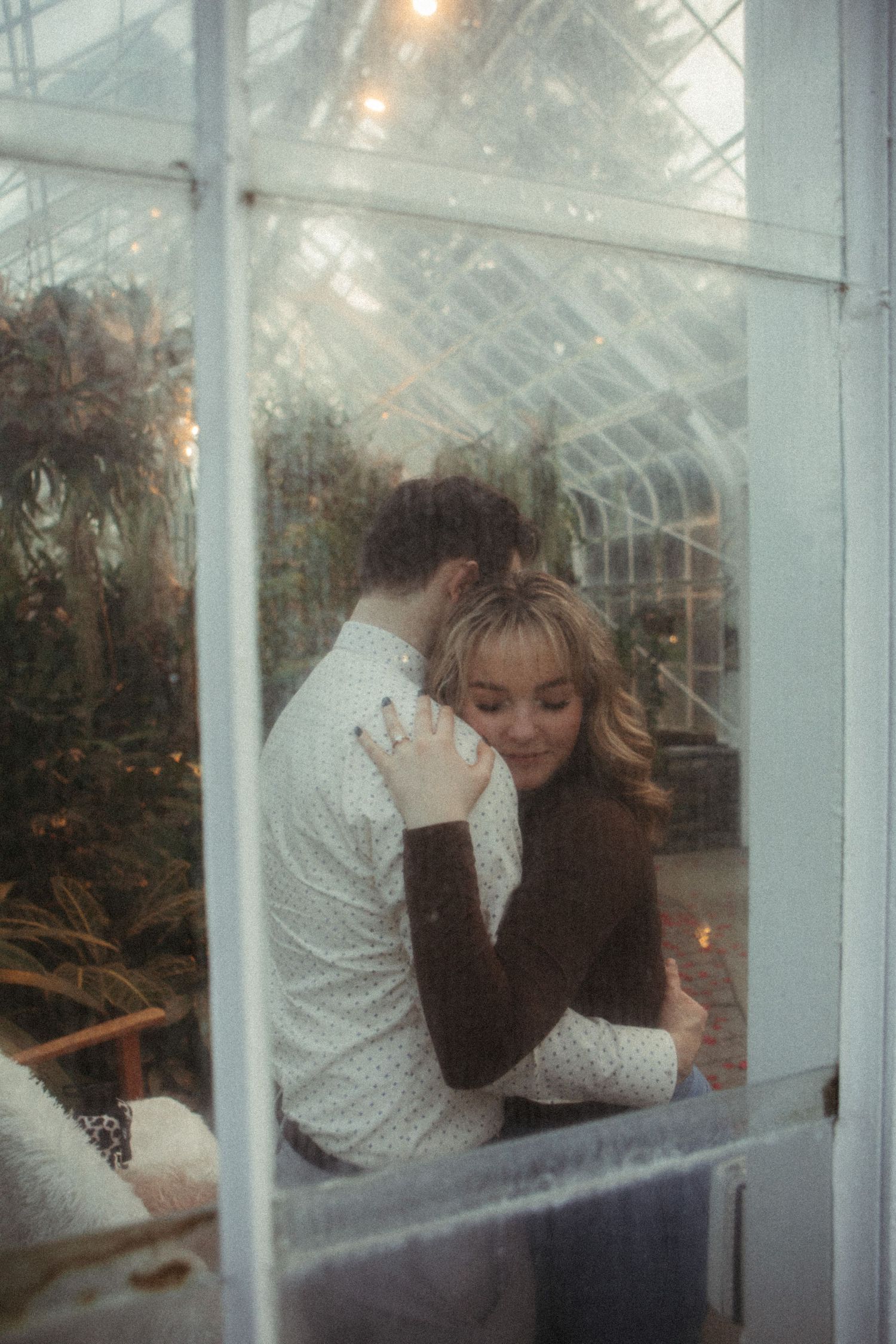 Couple embracing romantically inside a greenhouse with fairy lights and lush green plants visible through a white window frame.