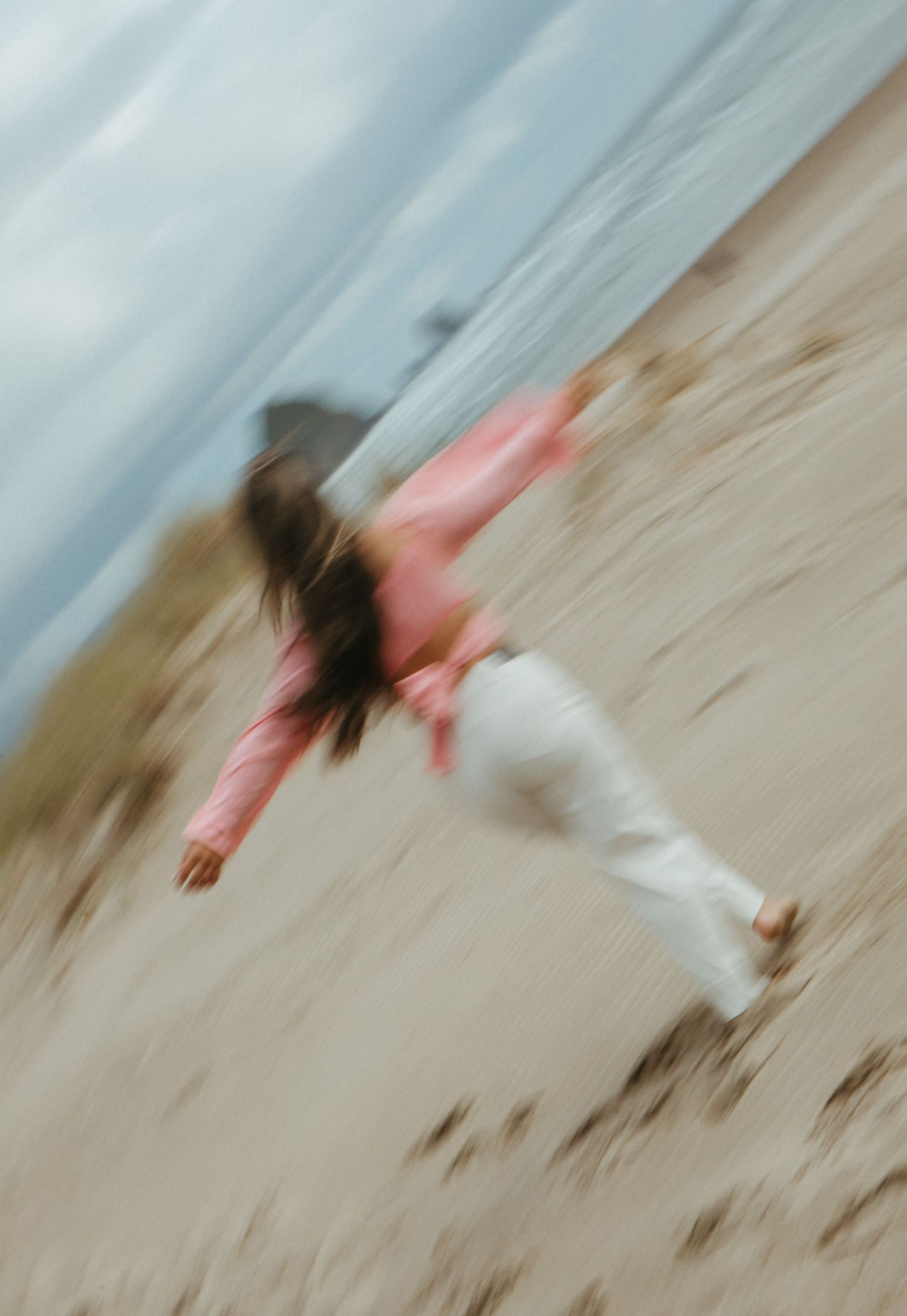 Motion-blurred figure in pink top and white pants leaping on a beach with cloudy sky in background.