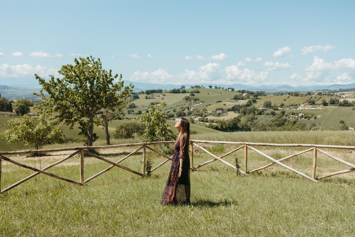Woman in floral dress standing by rustic wooden fence overlooking rolling Tuscan hills on a sunny day.