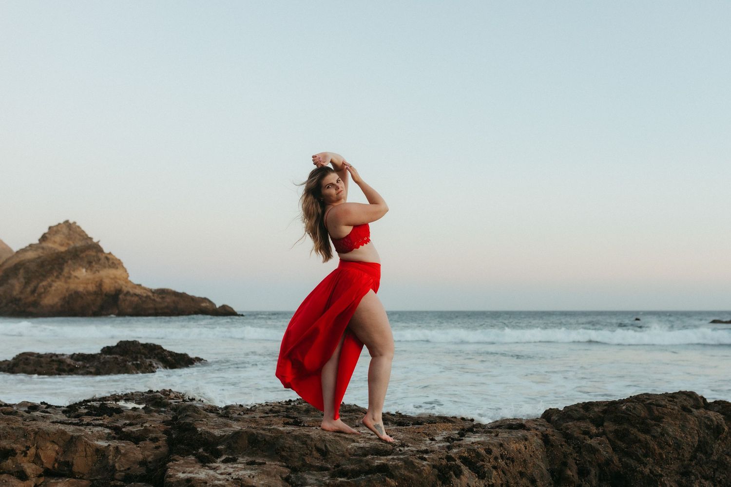 A red dress flows dramatically against the ocean backdrop as waves crash on rocky shore during golden hour.