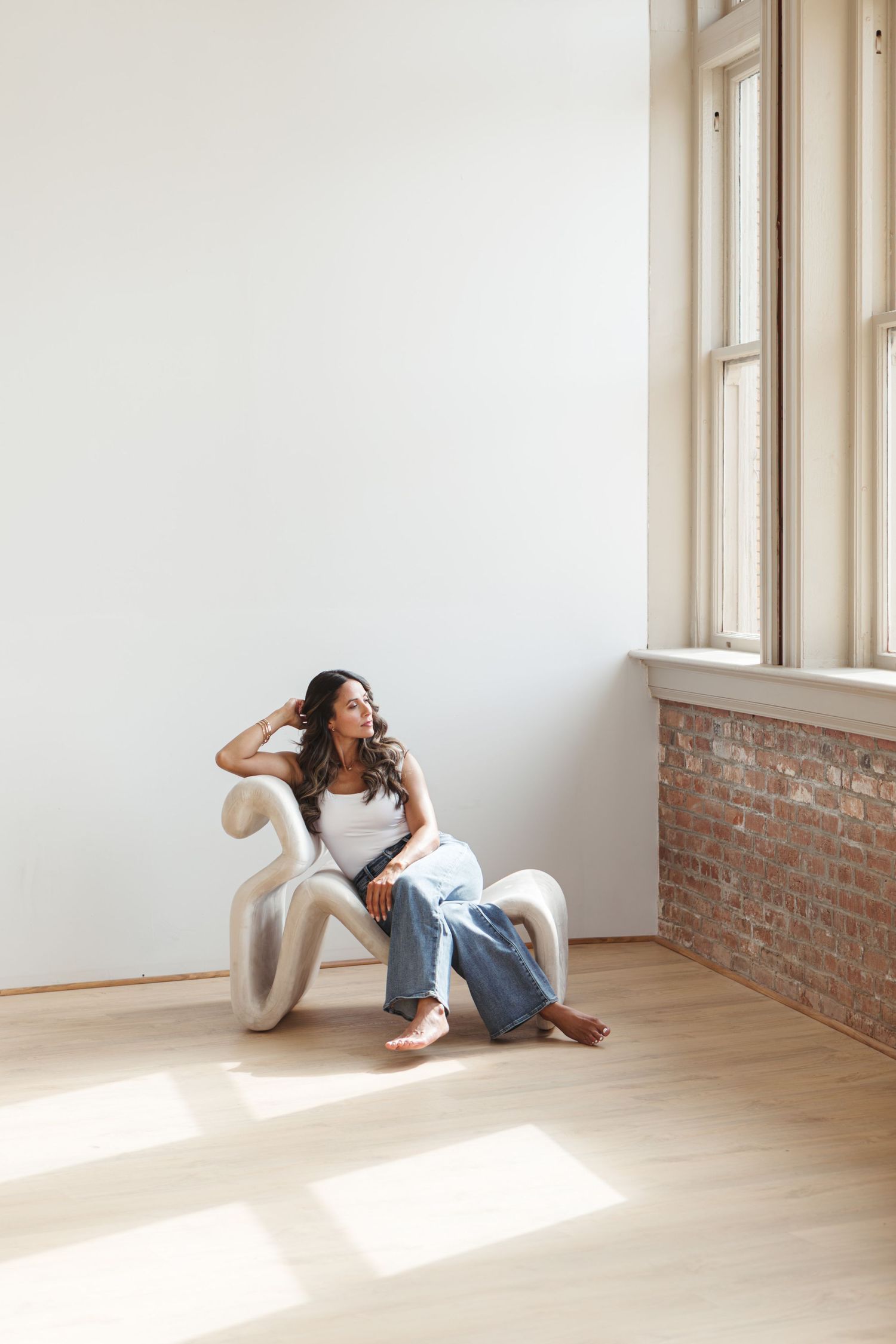 Woman with long wavy hair relaxes in a sculptural cream chair beside a sunlit window and exposed brick wall in a bright minimalist studio.