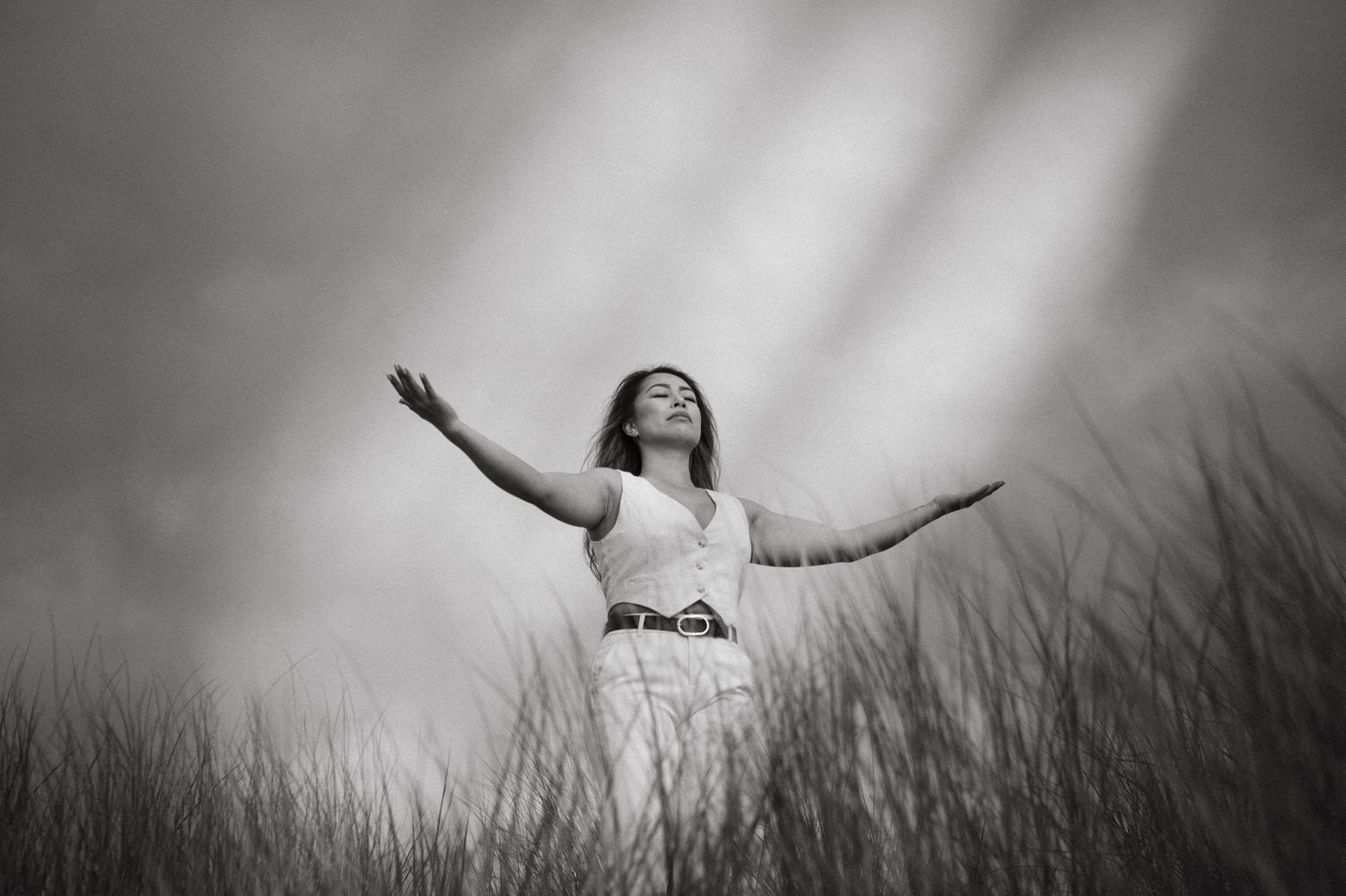 A person stands with outstretched arms in a field of tall grass against a moody black and white sky.