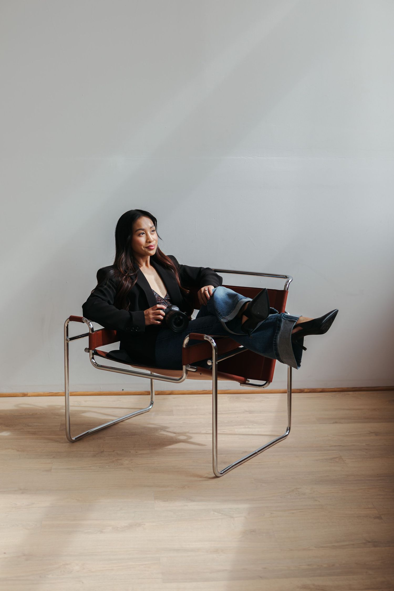 Woman in a black blazer and jeans lounging in a mid-century chrome and leather Wassily chair against a plain wall.