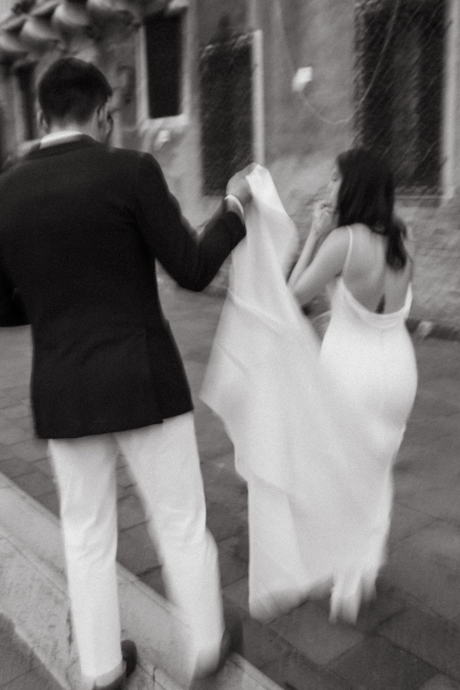 Blurred black and white photo of a bride and groom walking hand in hand through cobblestone streets.