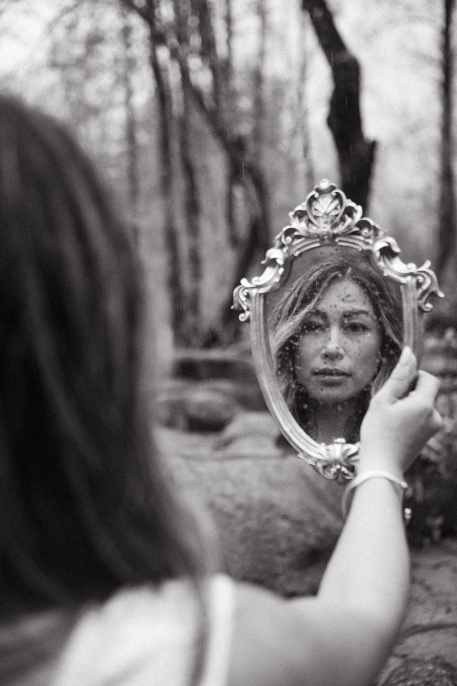 A hand holds an ornate vintage mirror reflecting a blurred forest scene in black and white.