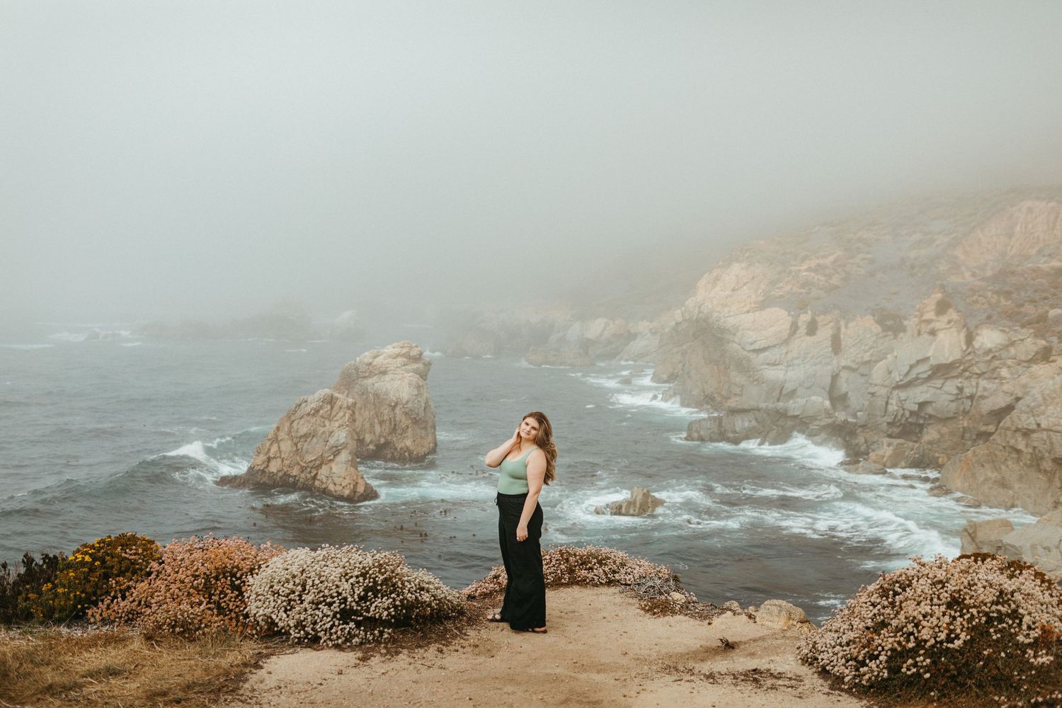 Foggy coastal landscape with rugged cliffs and crashing waves along the California coastline during golden hour.