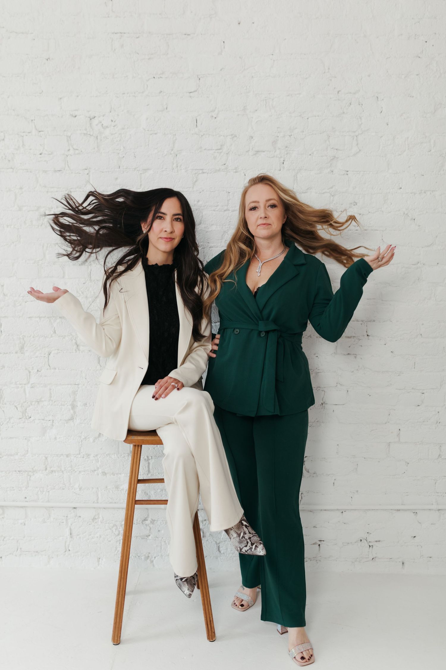 Two models in contrasting white and dark green suits pose dynamically against a white brick wall in a fashion photoshoot.