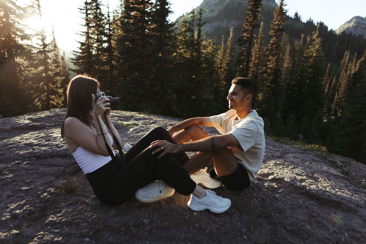 Two people sit together on a rocky mountain overlook during golden hour surrounded by evergreen trees and peaks.