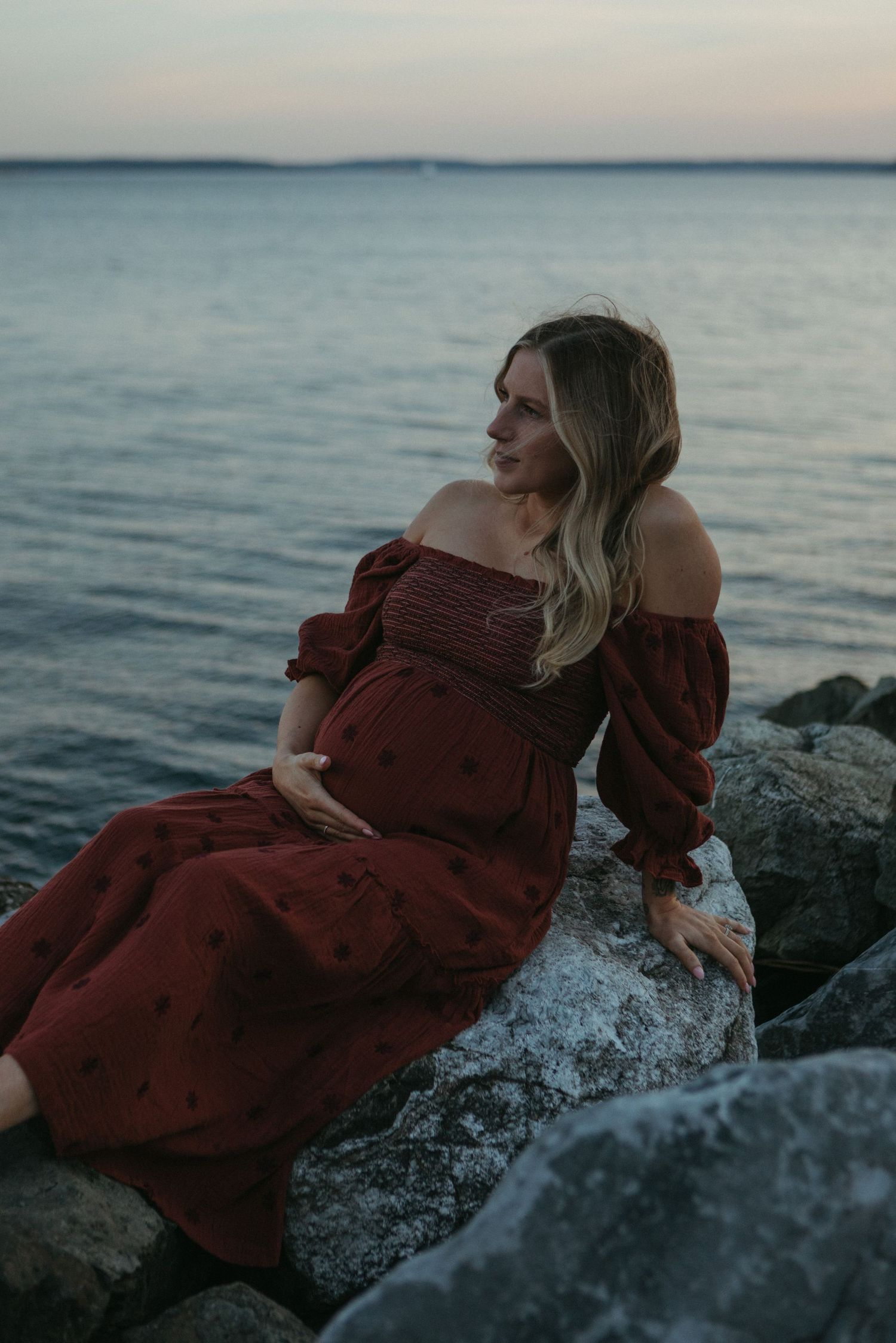 A person in a burgundy dress reclines on coastal rocks at sunset by the ocean.