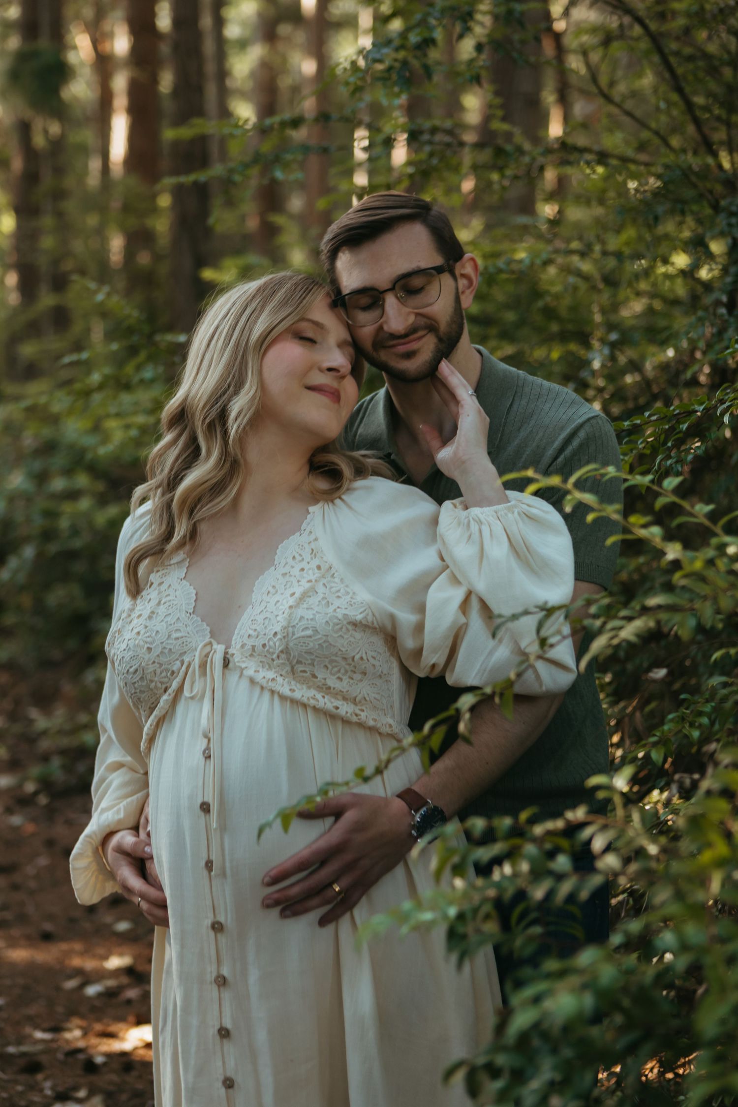 A romantic couple embraces in a sunlit forest wearing vintage-inspired clothing among green foliage.