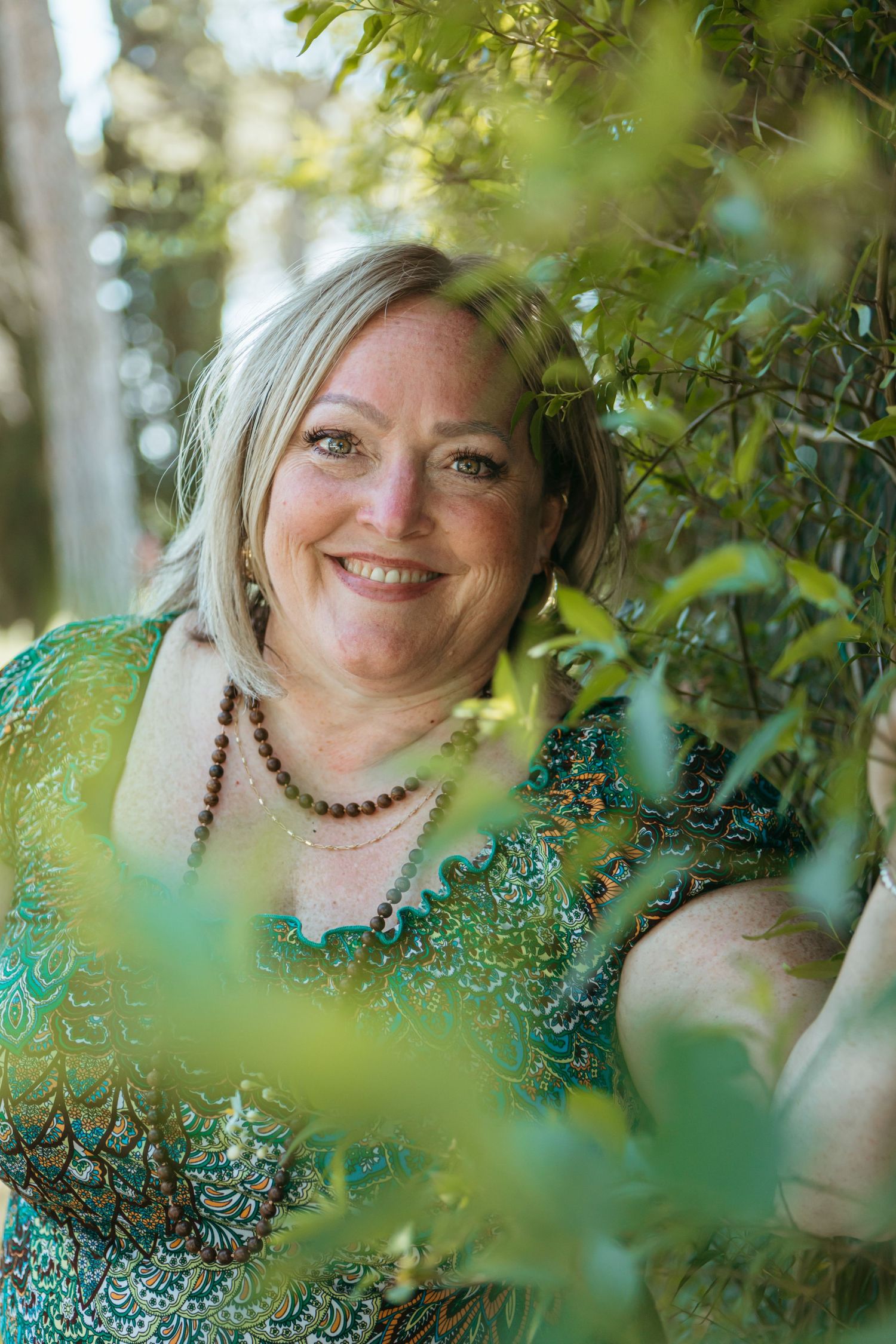 Smiling middle-aged woman with blonde hair wearing a green patterned dress and beaded necklace, surrounded by lush green foliage.