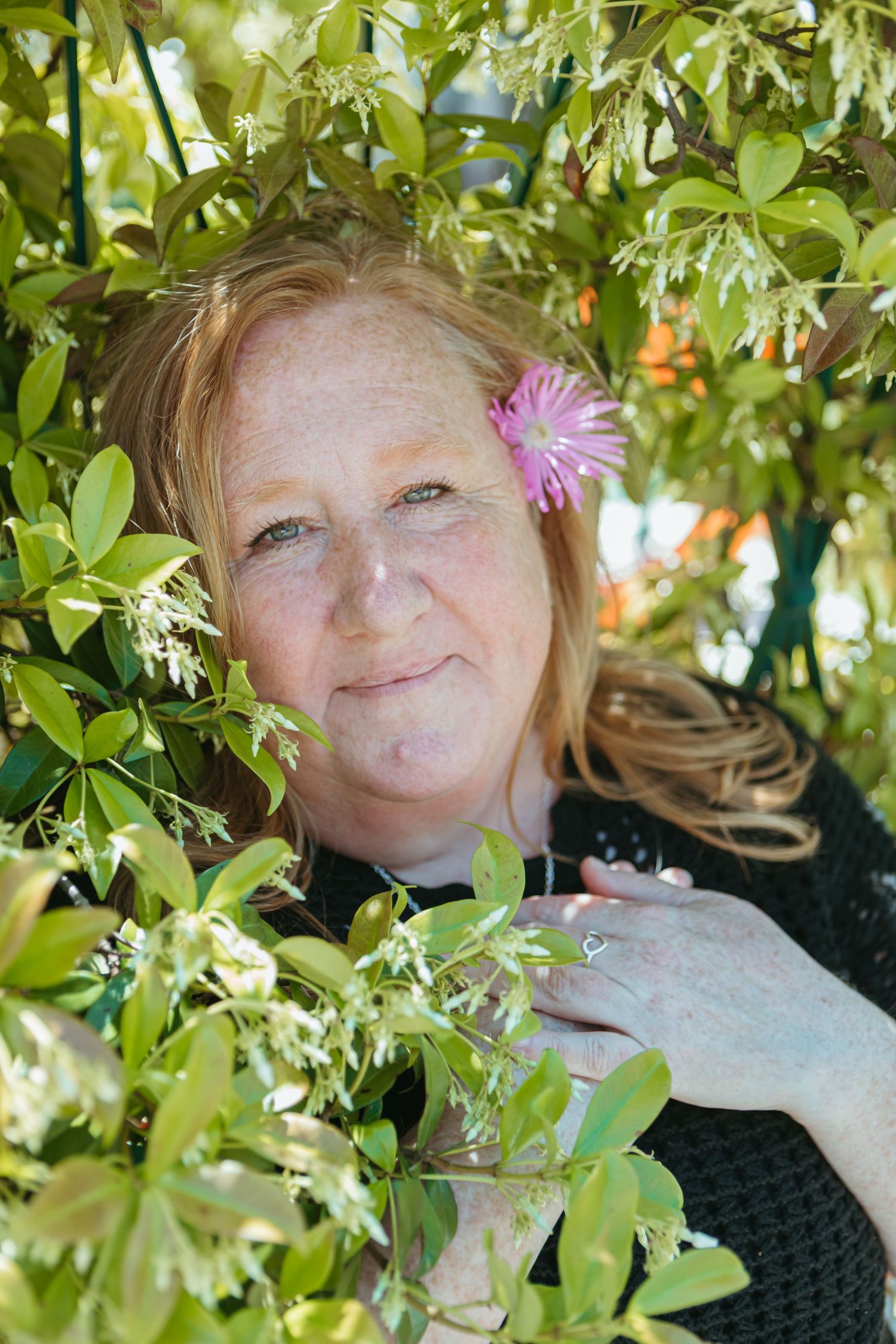 Red-haired woman with freckles and pink flower in hair smiling among lush green garden foliage.