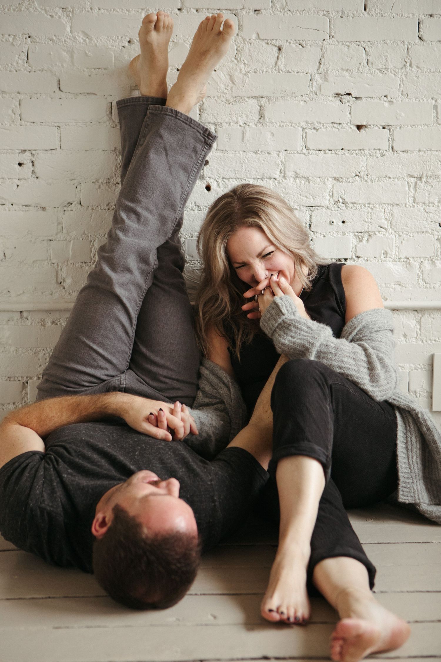 Laughing couple relaxing on floor against white brick wall, woman sitting up smiling while man lies with legs raised.