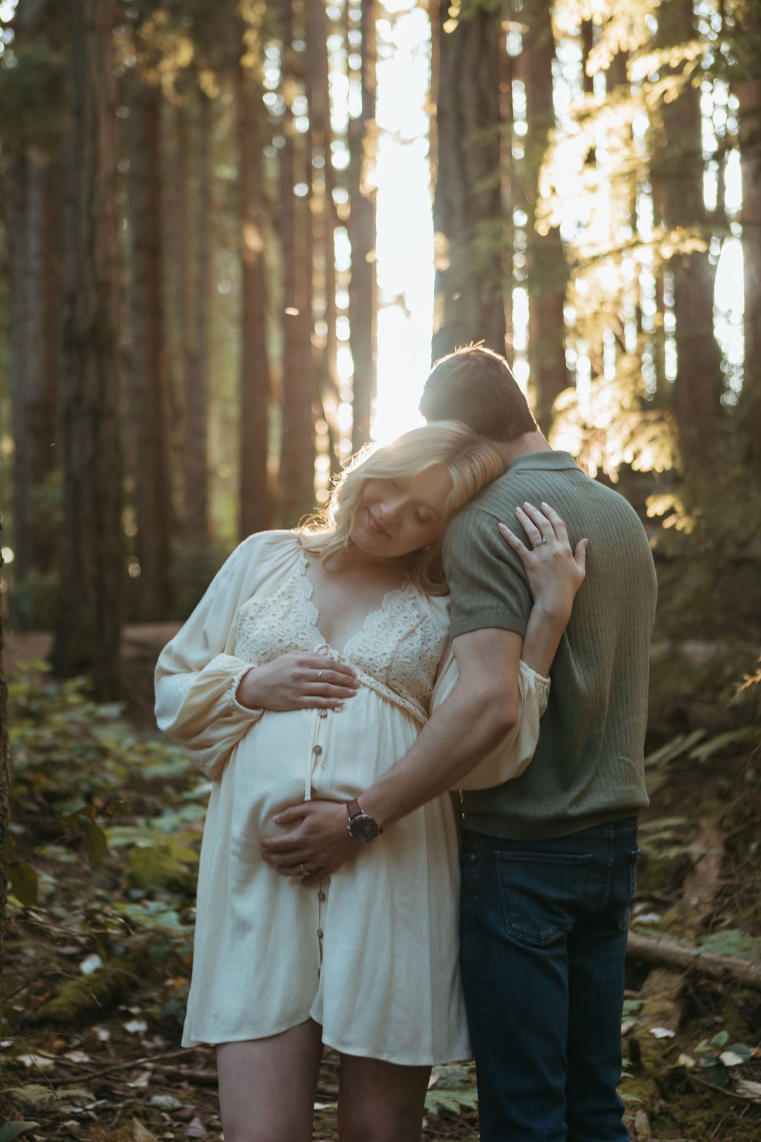 A couple embraces tenderly in a sunlit forest with tall trees and fallen leaves creating a romantic atmosphere.