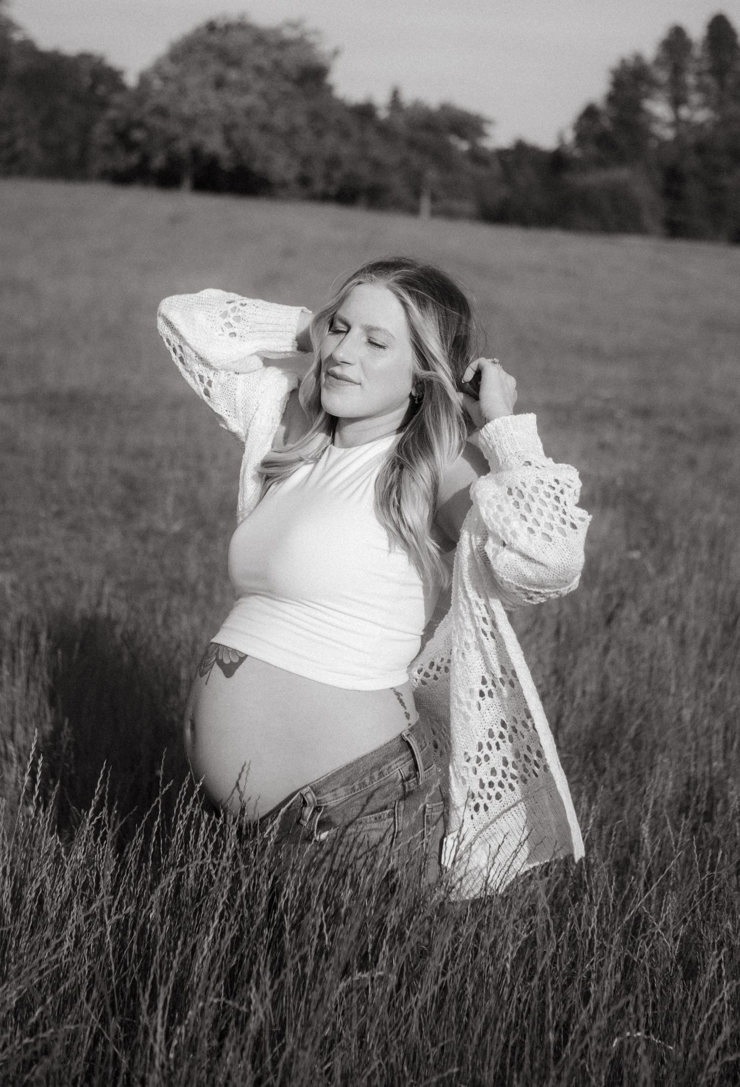 Expectant mother in a white dress relaxes in a sunny meadow with tall grass.