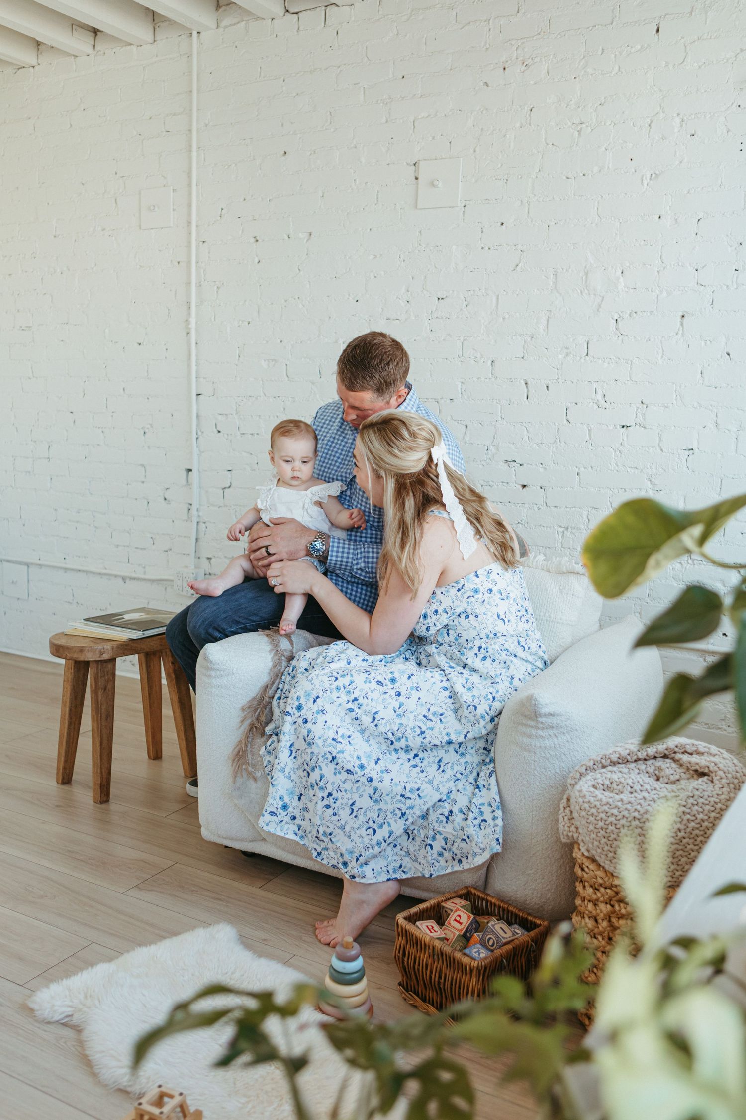 A young family with a baby sits together on a white sofa in a bright, modern living room with brick walls.