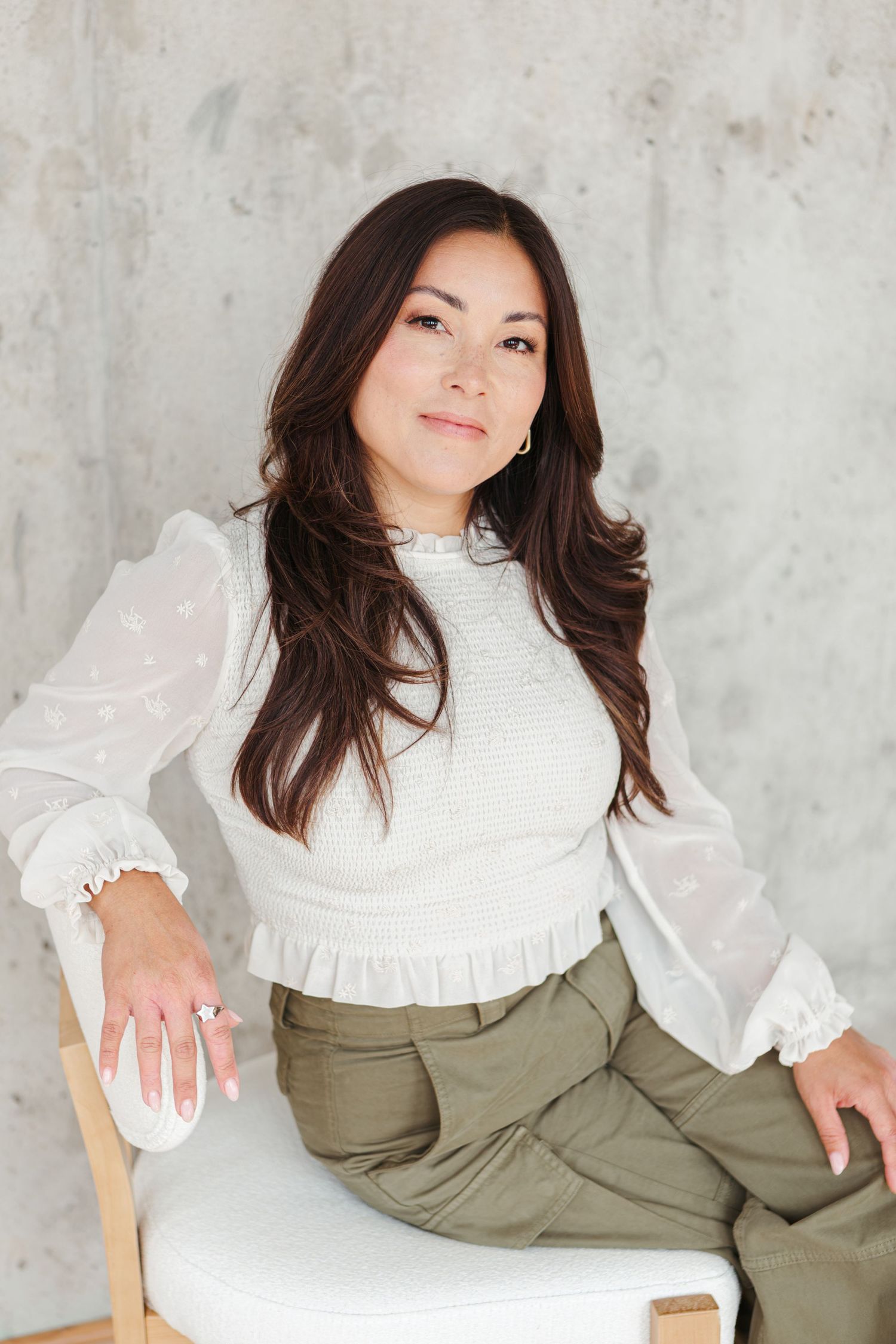 Woman with long dark wavy hair sits on a white cushioned chair in a cream ruffled blouse and olive green pants against a concrete wall.