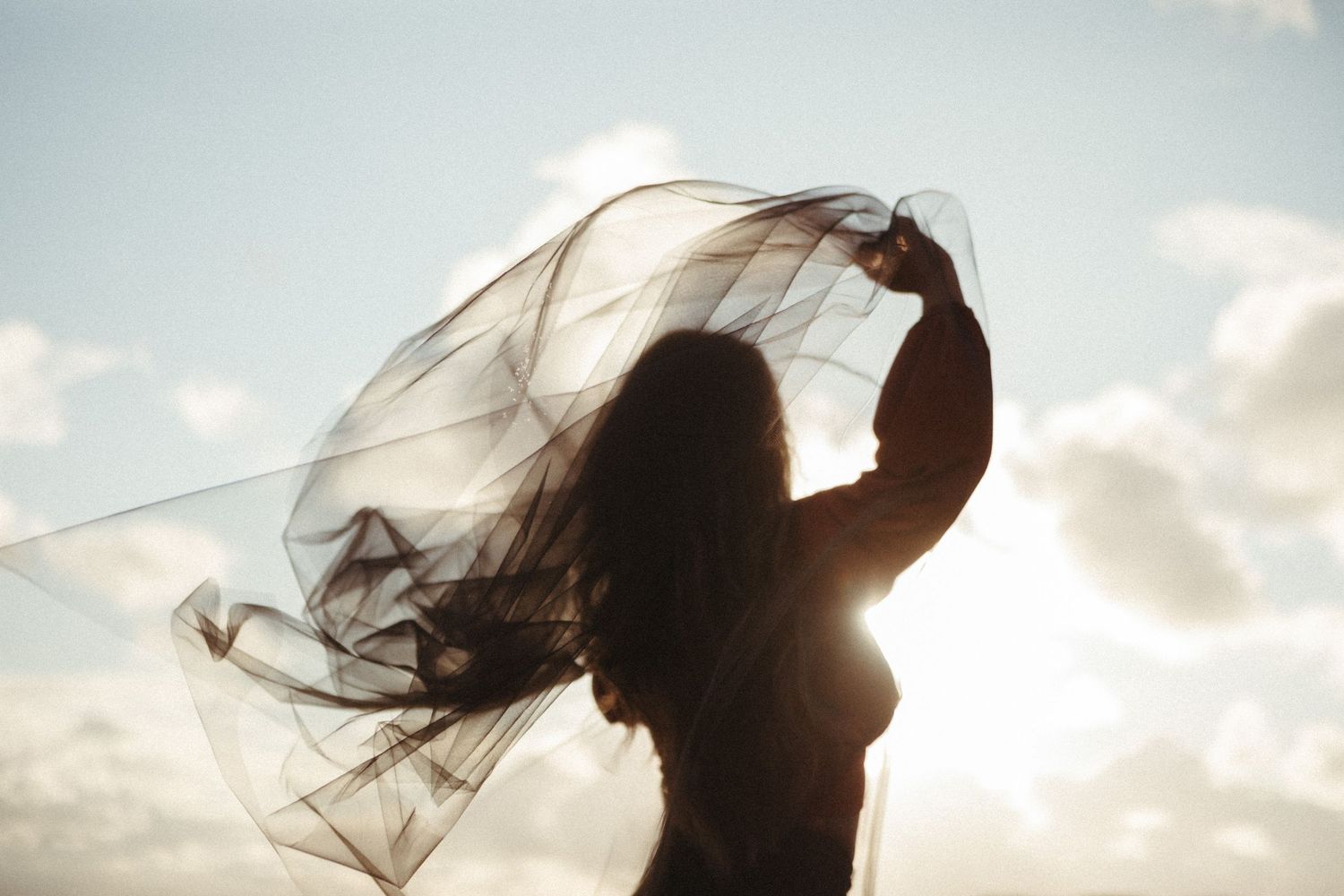 Silhouetted figures with flowing veils stand against a cloudy sky in a romantic wedding photo series.