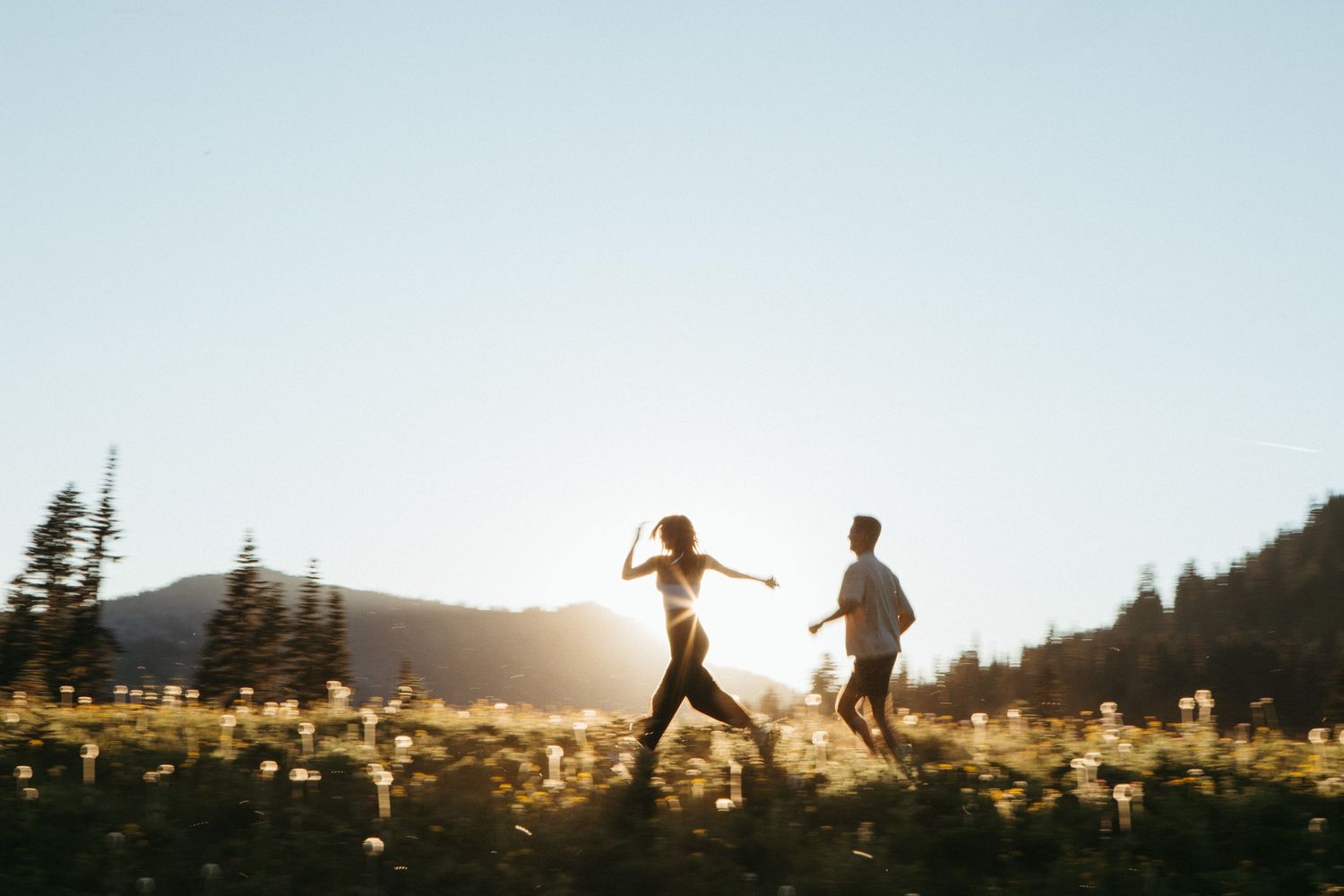 Silhouettes of people dancing and jumping at sunset in a mountain landscape with evergreen trees.