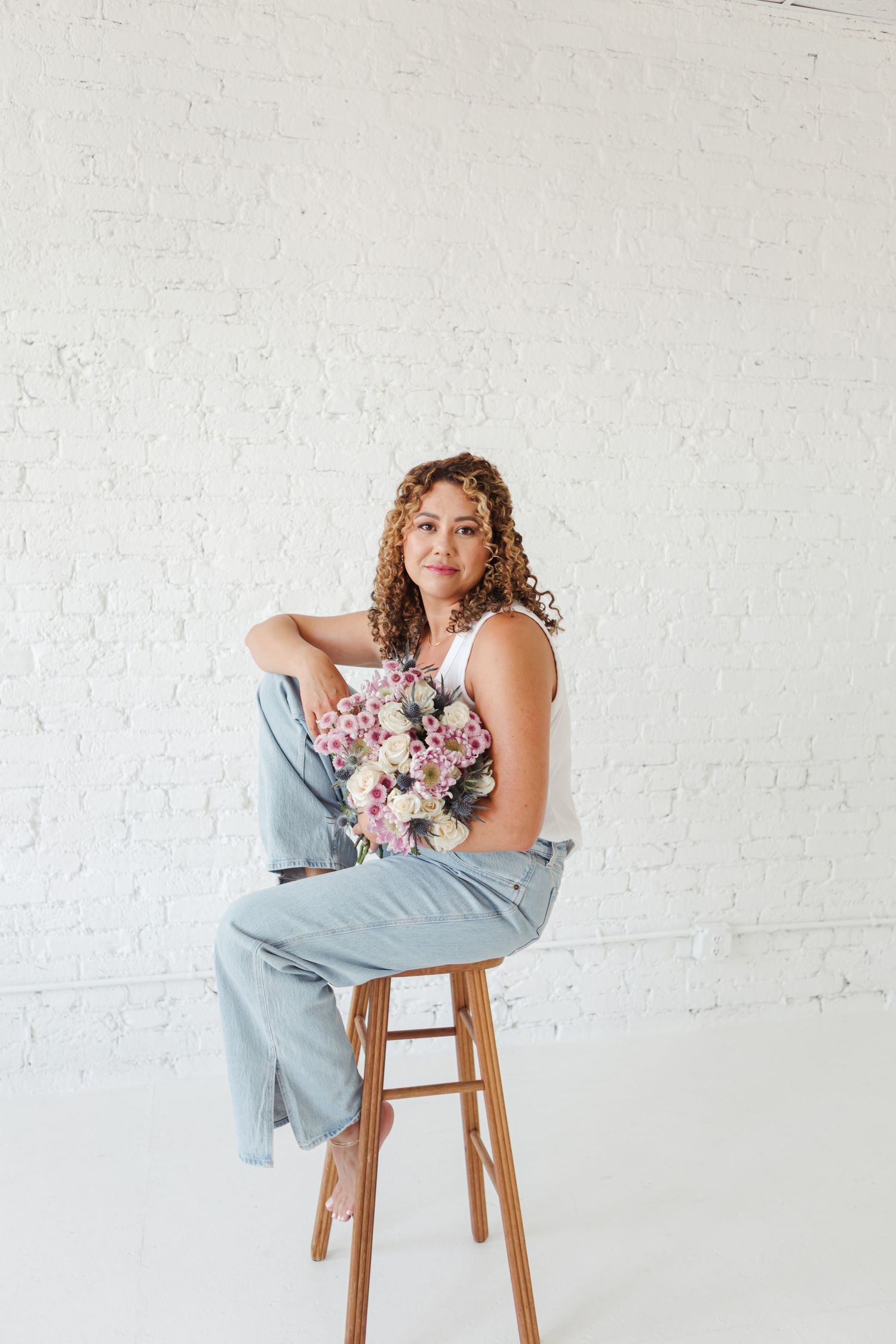 A figure in a light blue dress poses gracefully on a wooden chair with a pink floral bouquet against a white backdrop.